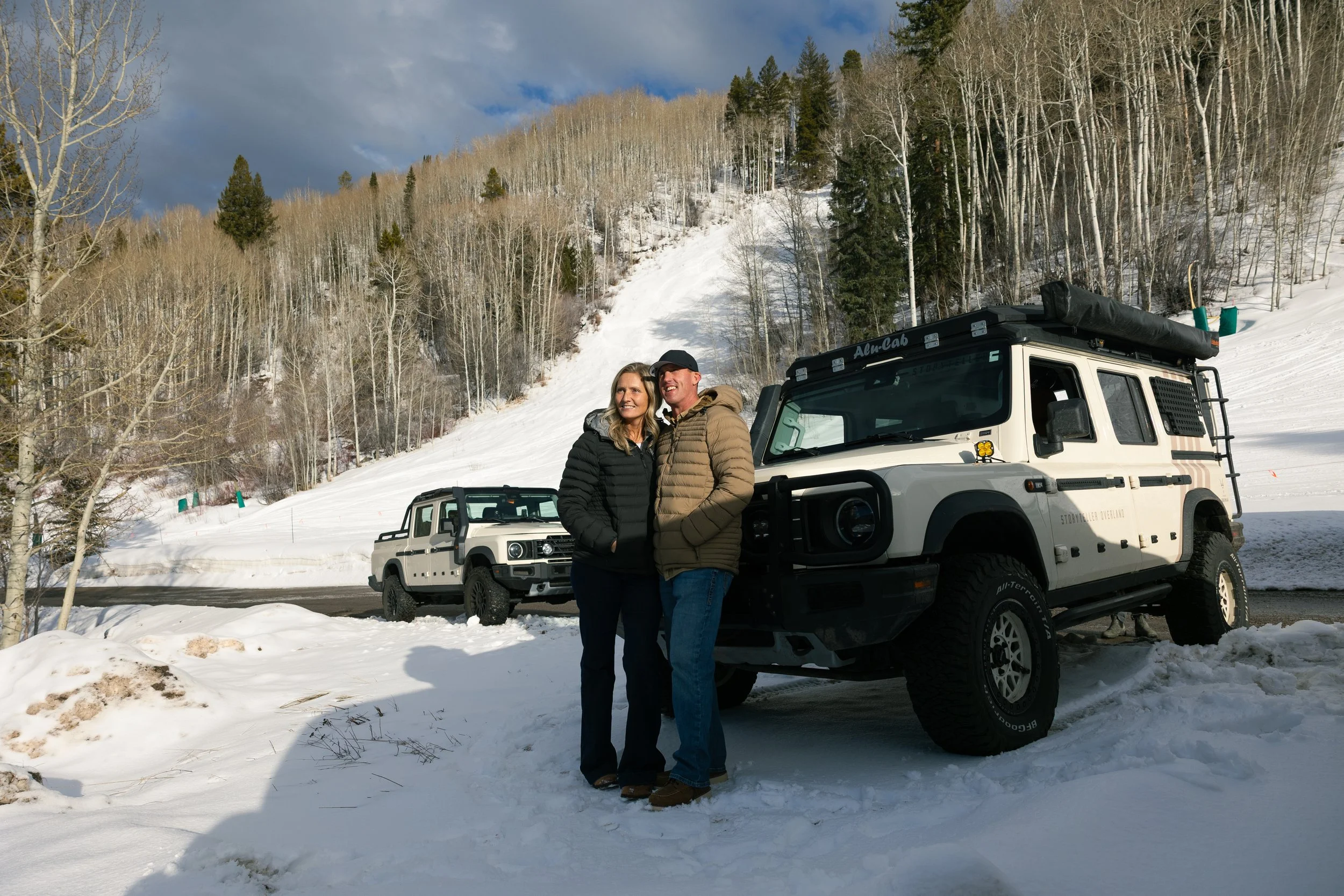 A couple stands smiling in winter clothing in front of two white off-road vehicles parked on snow, with snow-covered mountains and leafless trees in the background.