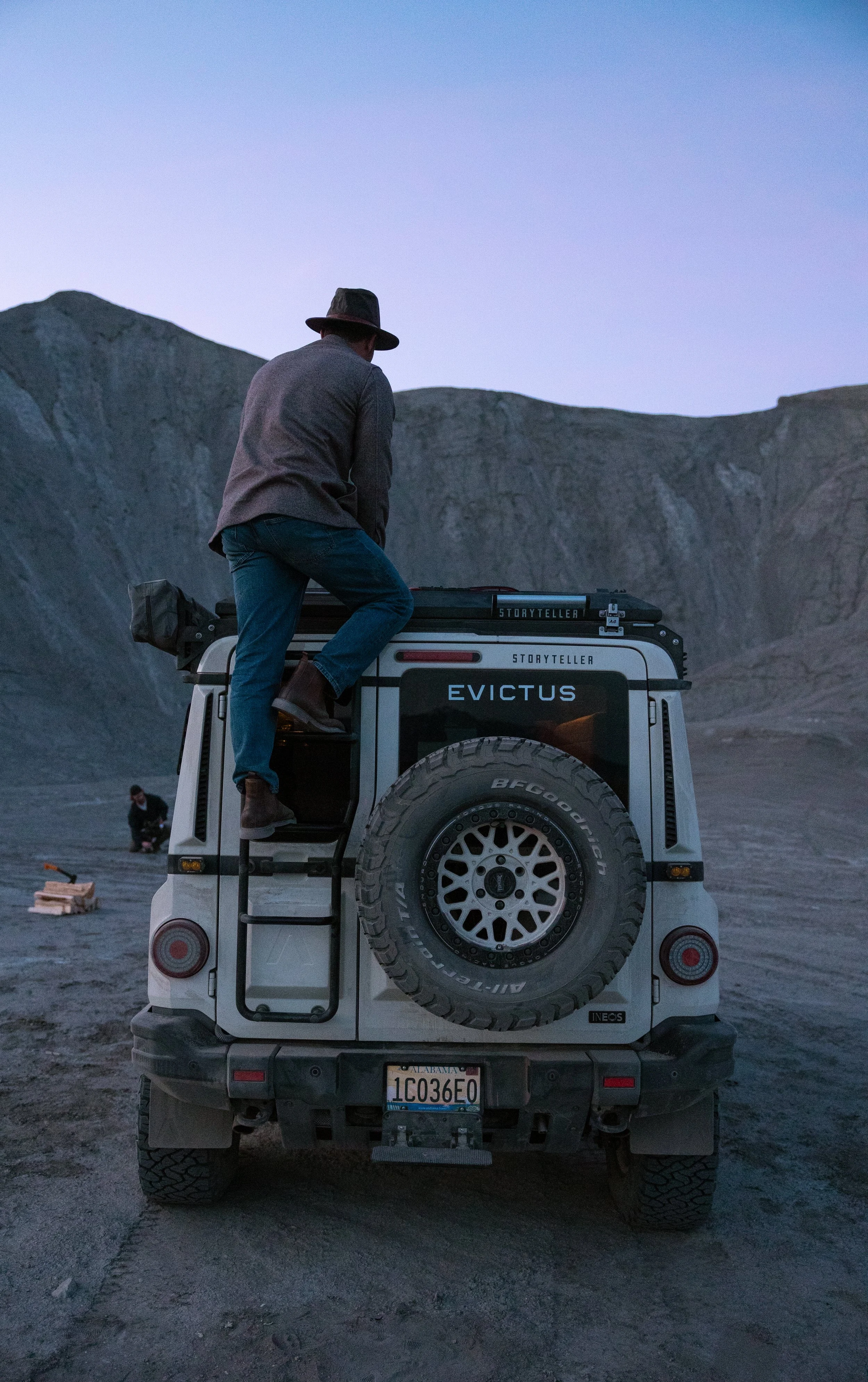 A person wearing a hat and jacket climbing on top of a Jeep vehicle in a rocky, outdoor landscape at dusk.