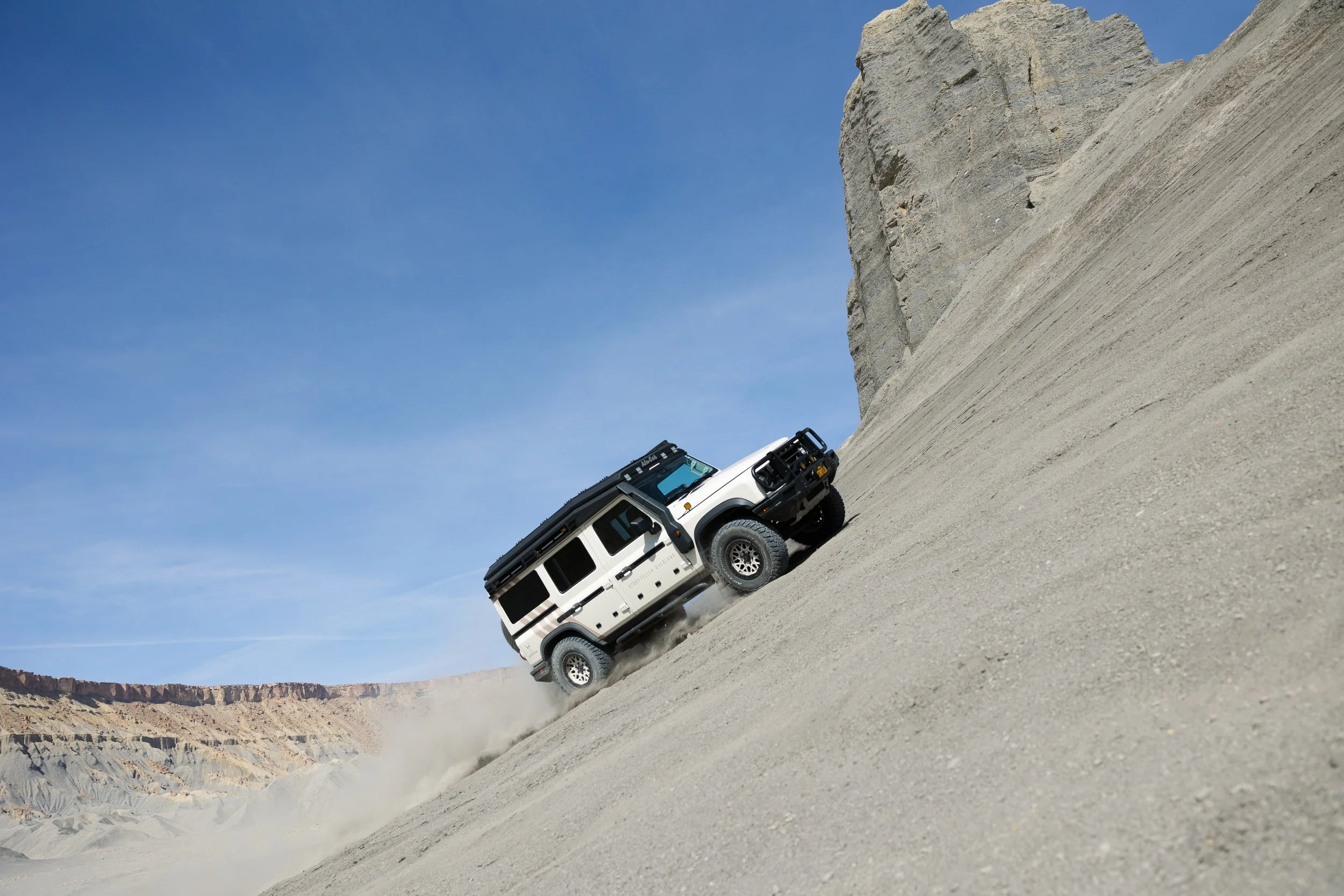 A white off-road vehicle ascending a steep sandy hill in a desert landscape with a clear blue sky.
