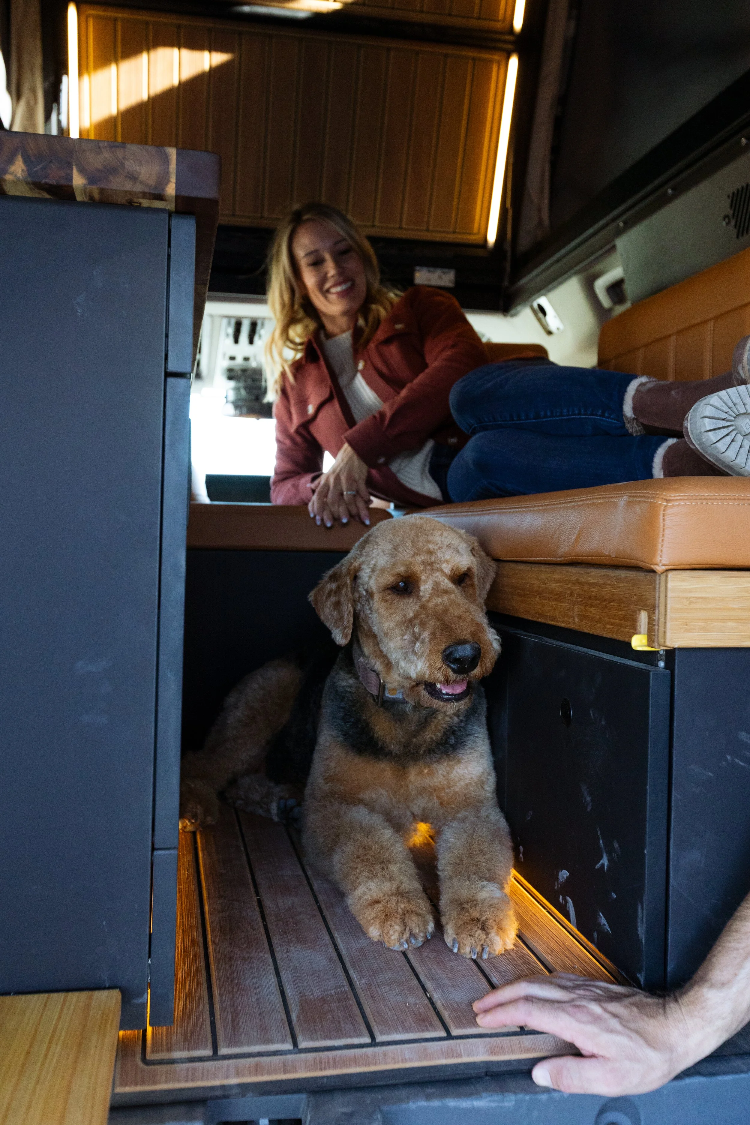 A woman smiling in a camper van with a brown and black dog lying underneath the bench seat.