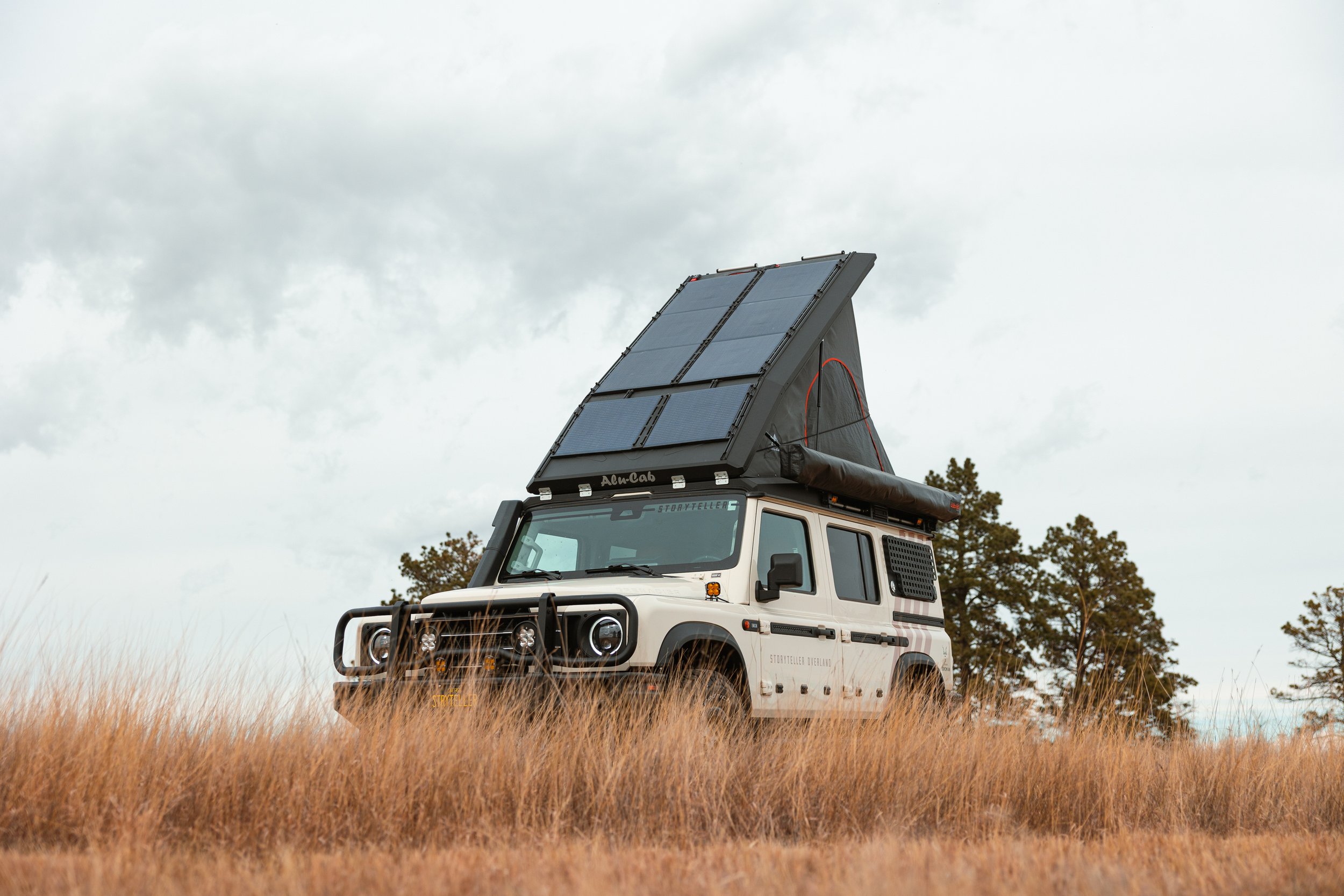 A white off-road vehicle with a solar panel rooftop tent, parked in a grassy field with trees in the background under cloudy skies.