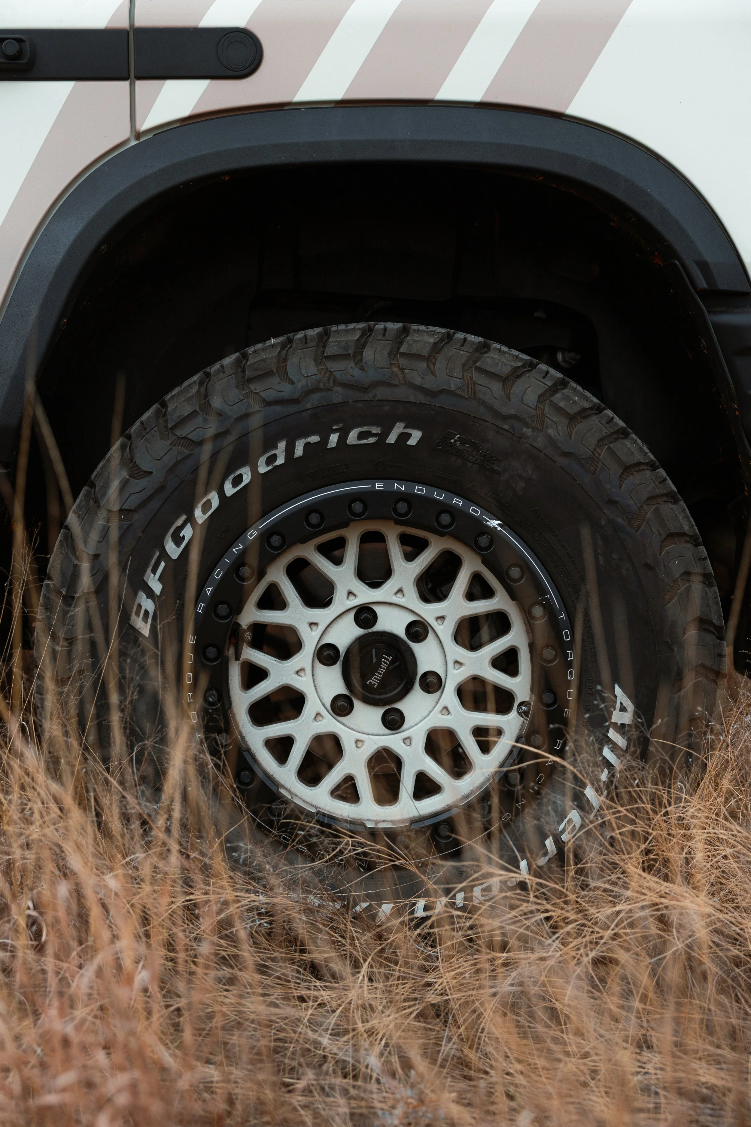 Close-up of a rugged off-road tire with a white alloy wheel, parked among tall dry grass, part of a white vehicle with black accents.
