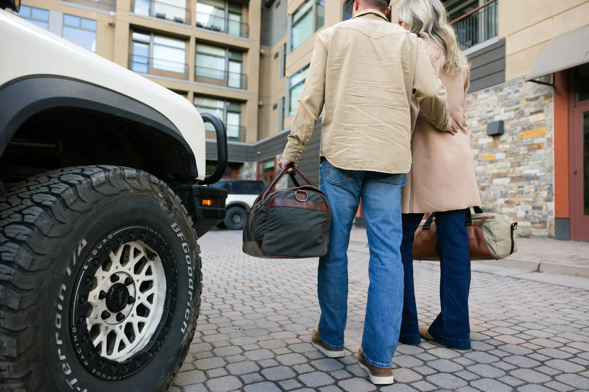 Two people standing outside near a large vehicle, possibly a truck or SUV, with luggage. They are dressed casually and appear to be waiting or preparing to leave.