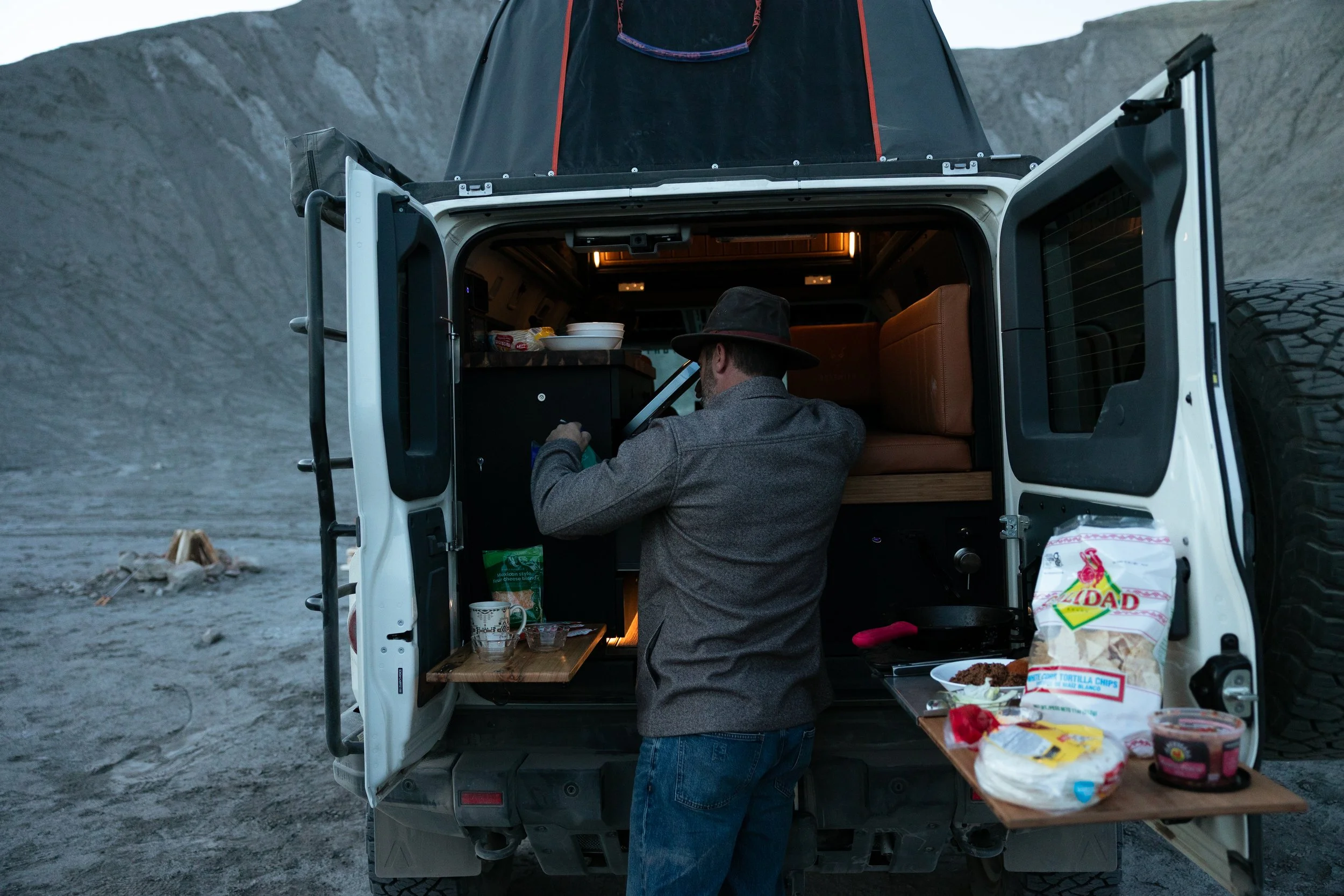 A man in a gray jacket and a dark hat stands at the back of an off-road vehicle, preparing food inside the vehicle. The vehicle's tailgate is lowered, revealing a compact kitchen setup with food and drinks, and some items placed on a fold-out table. 