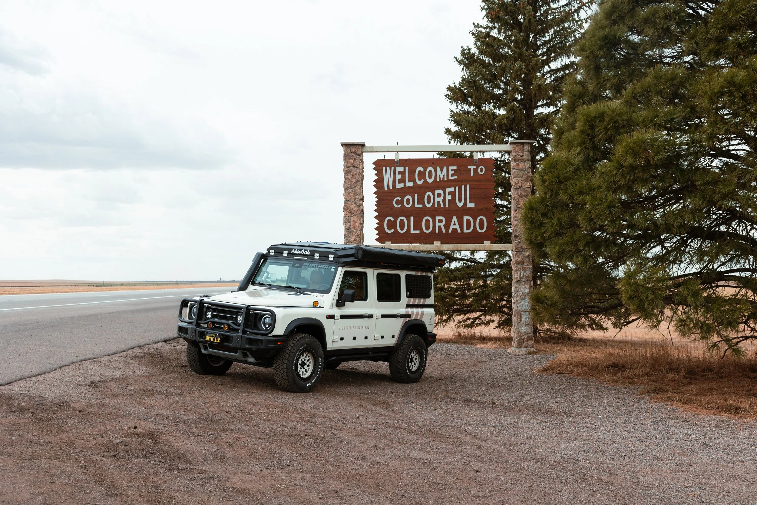A white off-road vehicle parked near a roadside sign that says "Welcome to Colorful Colorado" on a partly cloudy day.