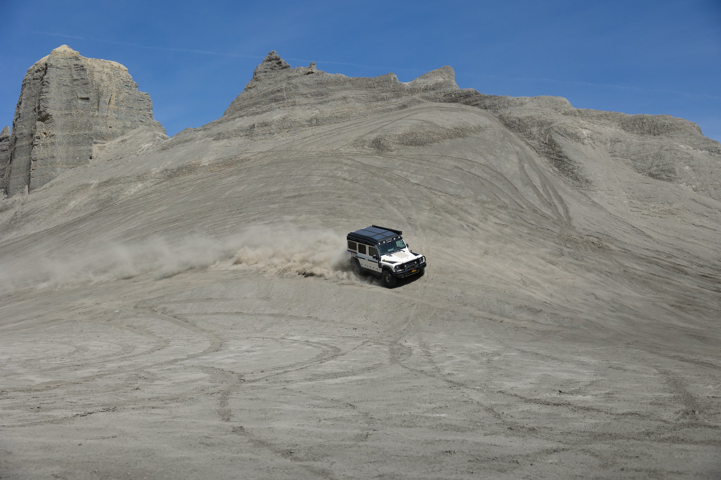 A white off-road vehicle driving on a sandy, desert-like landscape with mountains and cliffs in the background, leaving a trail of dust behind.