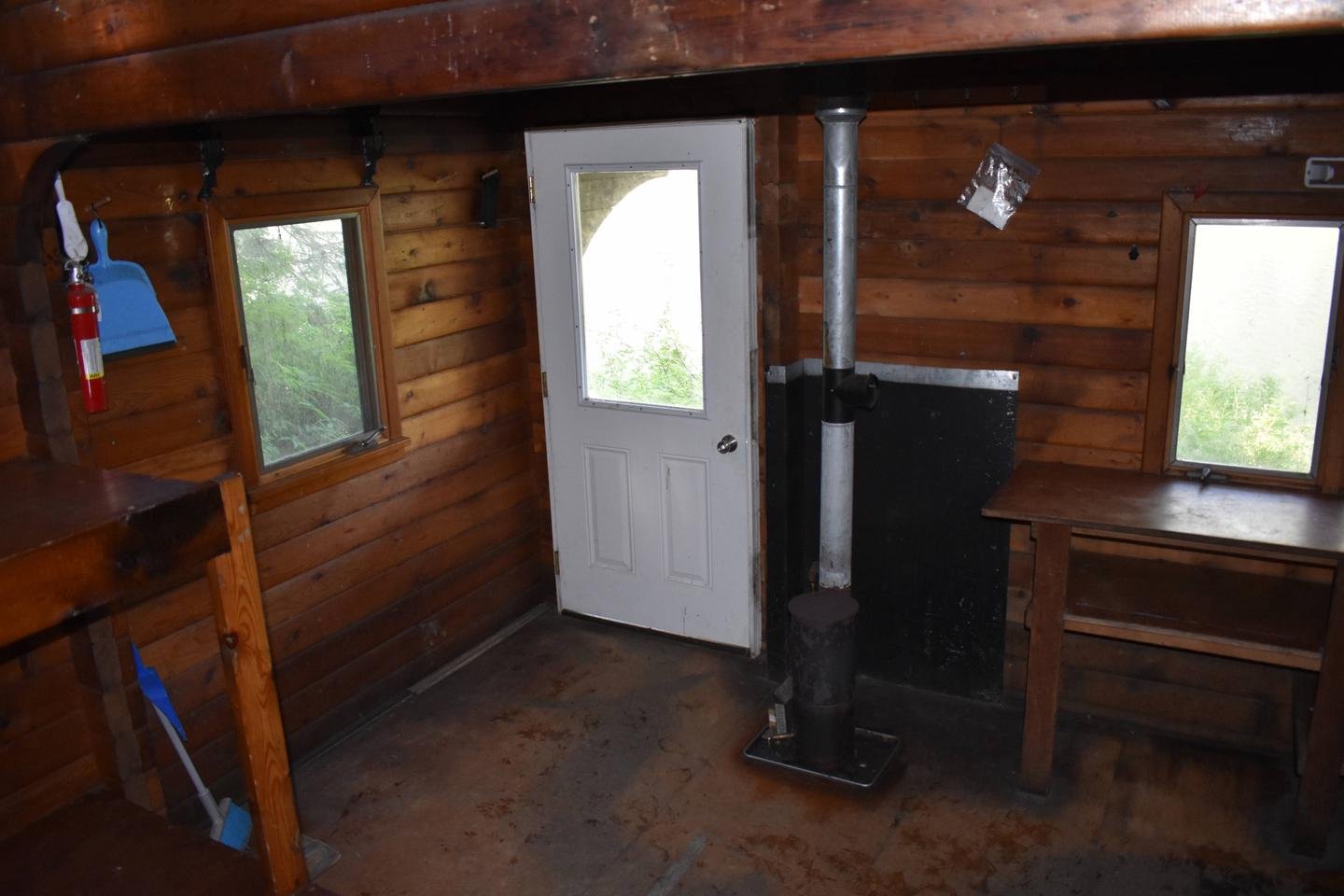 Interior of a wooden cabin with small windows, a white door with a window, a wood stove with a pipe, a workbench, shelves, and cleaning tools.