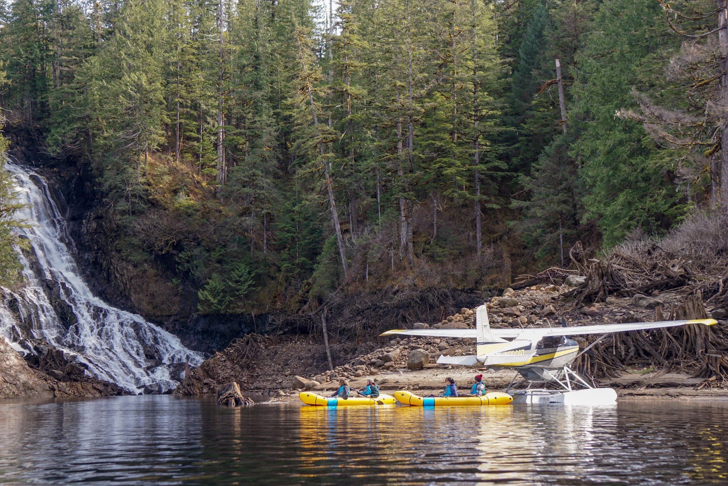 People rafting on a river next to a forested area with a waterfall, with a seaplane docked nearby.