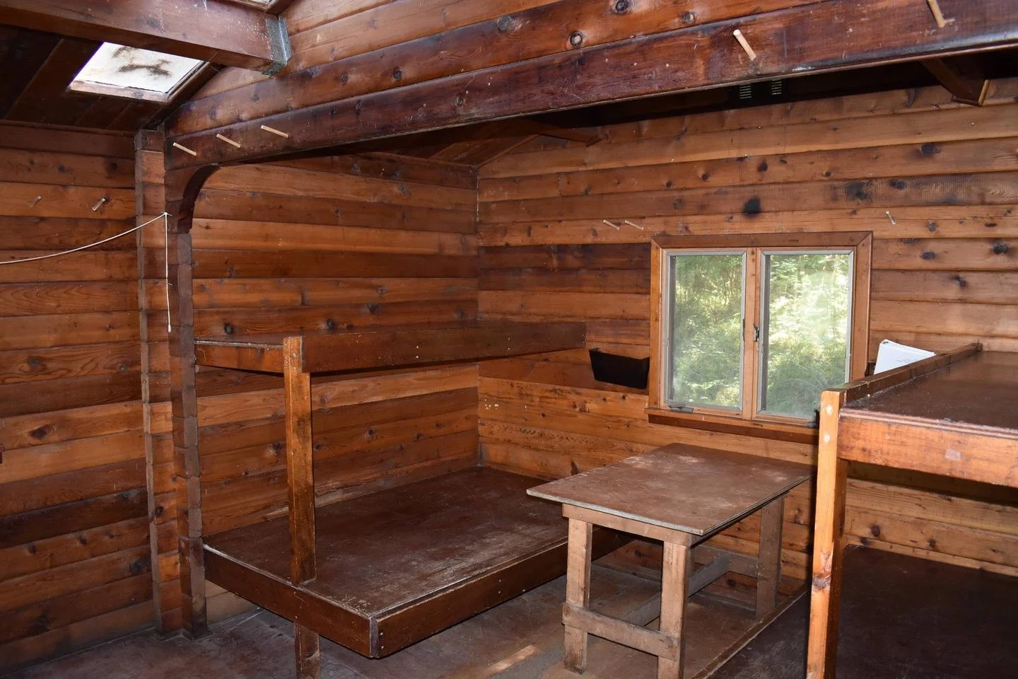 Interior of a rustic wooden cabin with log walls, a small window showing greenery outside, a loft bed, and a simple wooden table.
