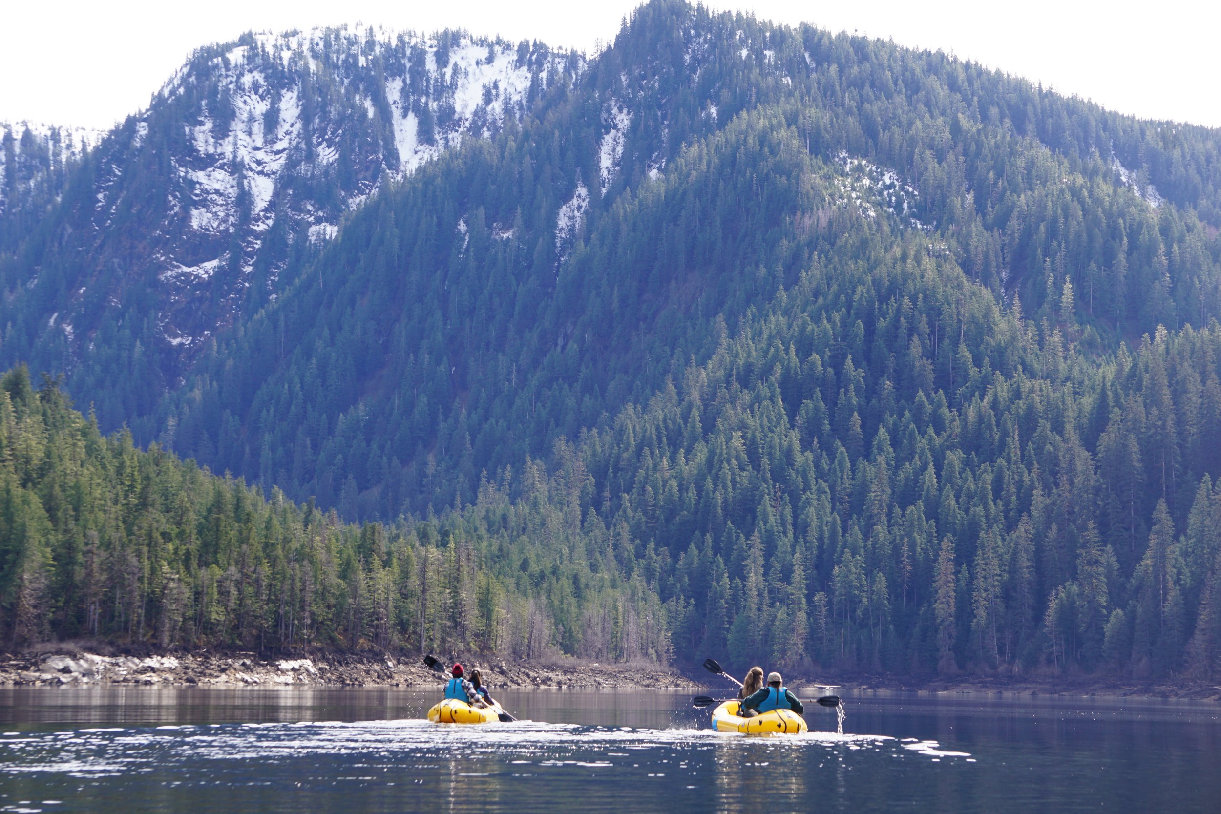 Two people in yellow kayaks paddling on a calm lake surrounded by dense evergreen forests, with a mountain in the background.