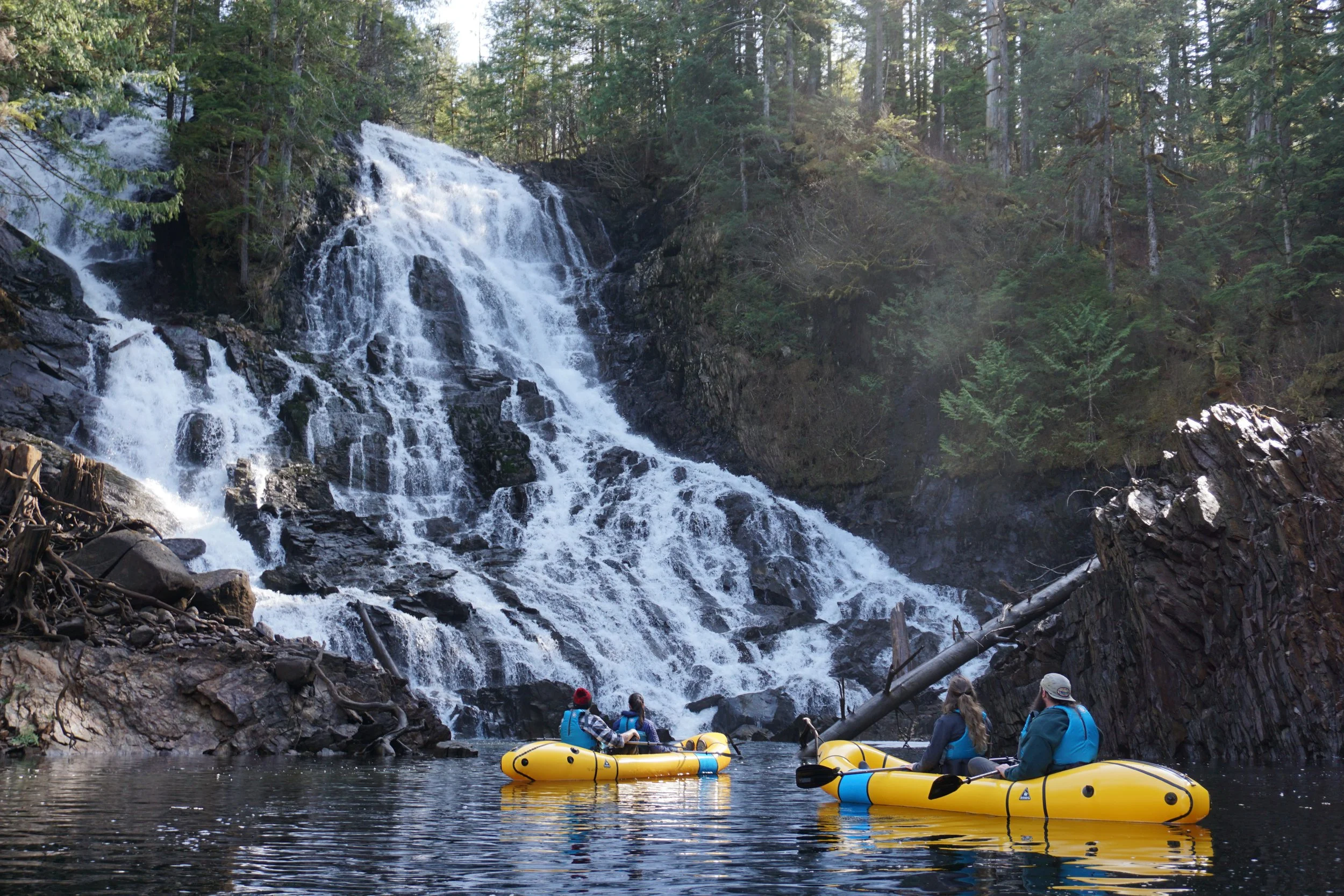 People kayaking in a river in front of a waterfall surrounded by trees.
