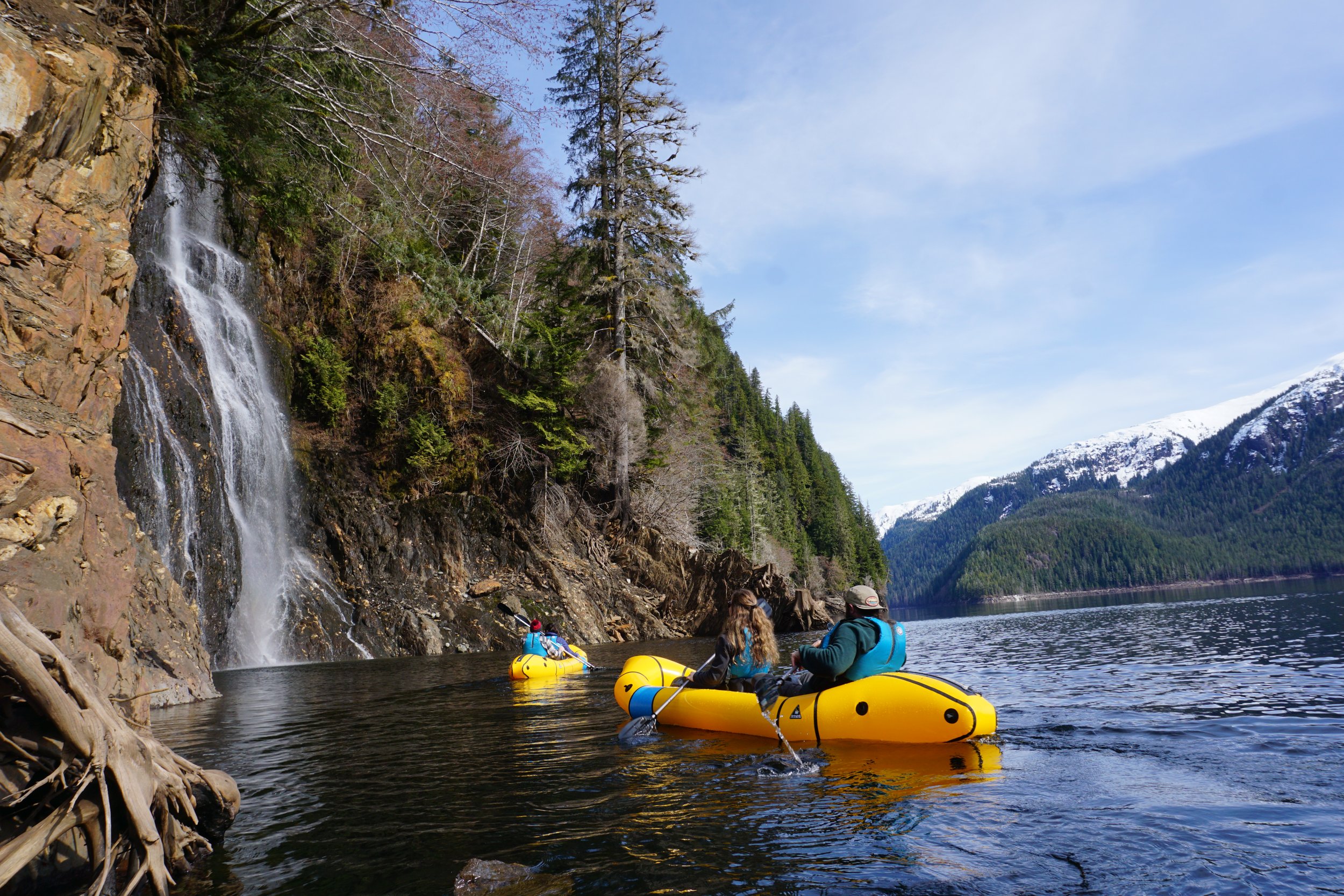 People kayaking on a river near a waterfall with mountainous, forested landscape and snow-capped mountains in the background.