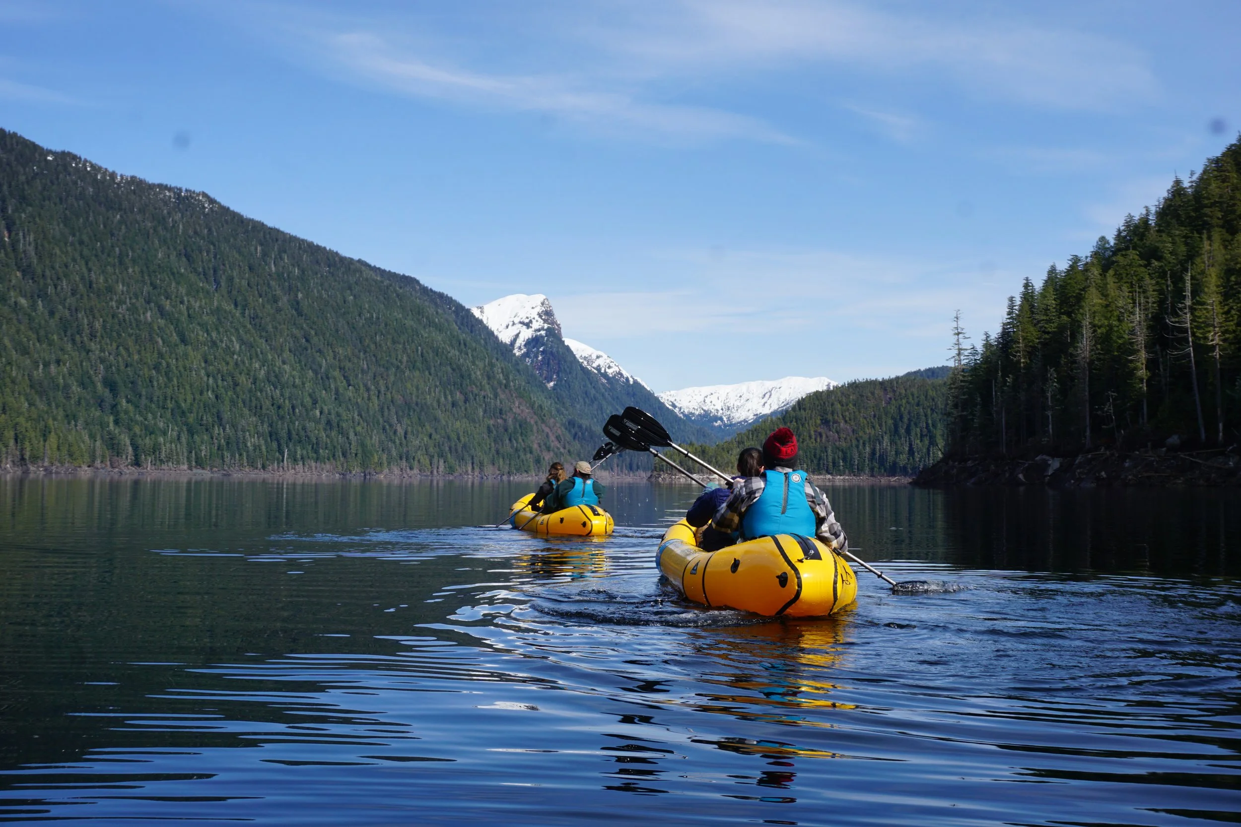 Two people in yellow kayaks paddling on a calm lake surrounded by green mountains with snow-capped peaks under a blue sky.