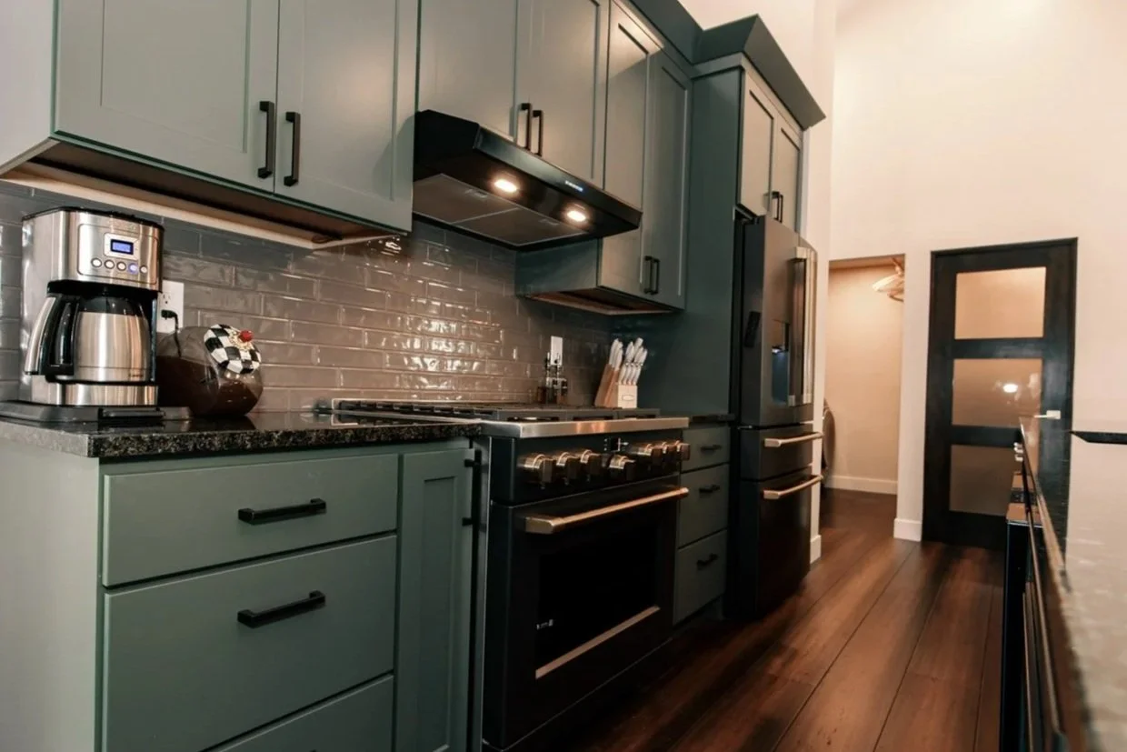 Modern kitchen with teal cabinets, black hardware, gray subway tile backsplash, black appliances including a refrigerator and a stove, a coffee maker, and a wooden knife block on the countertop.