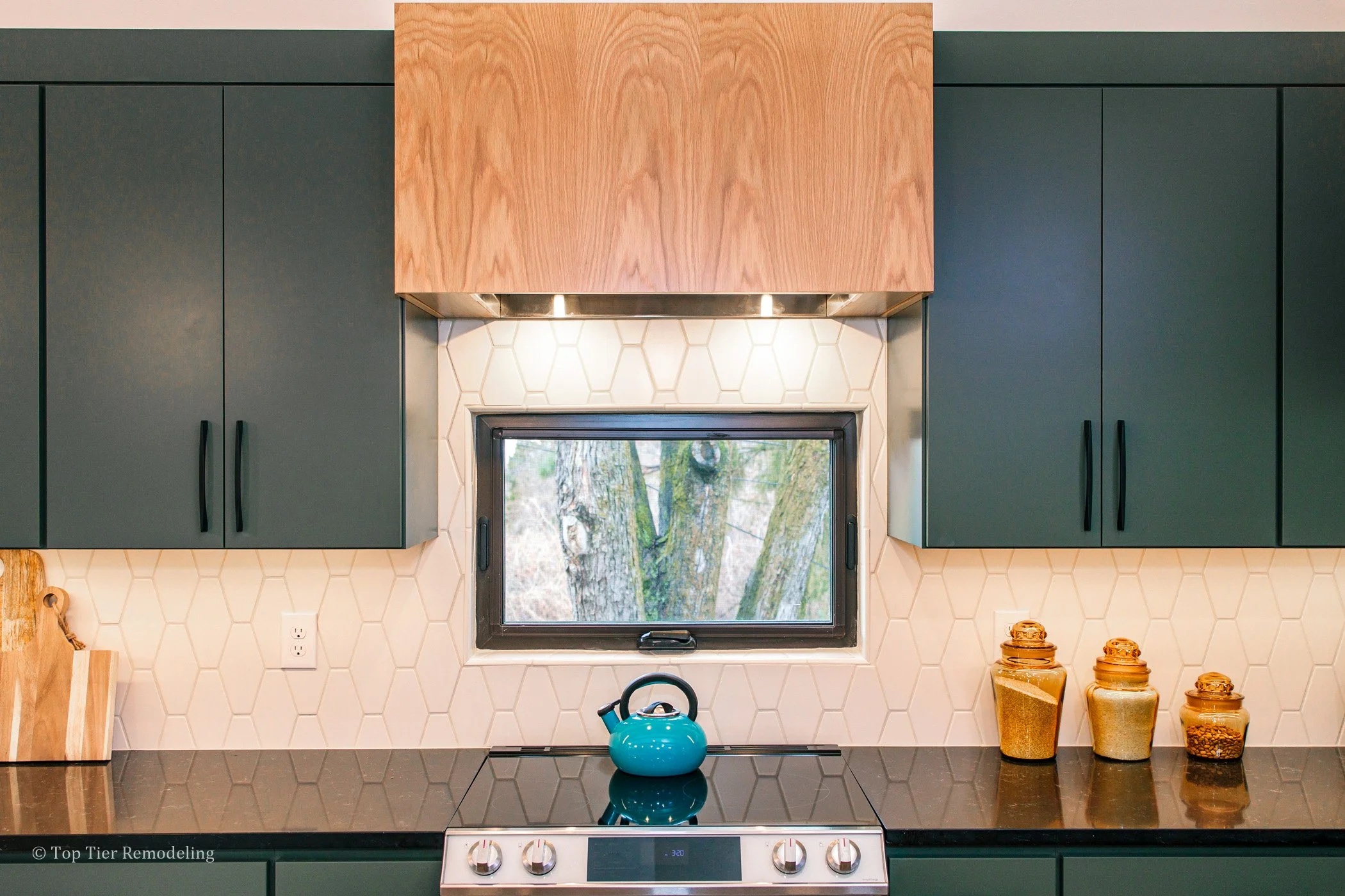 A modern kitchen with dark green cabinets, a black countertop, a backsplash with white honeycomb tiles, and a small window showing trees outside. There are some jars and a blue kettle on the countertop.