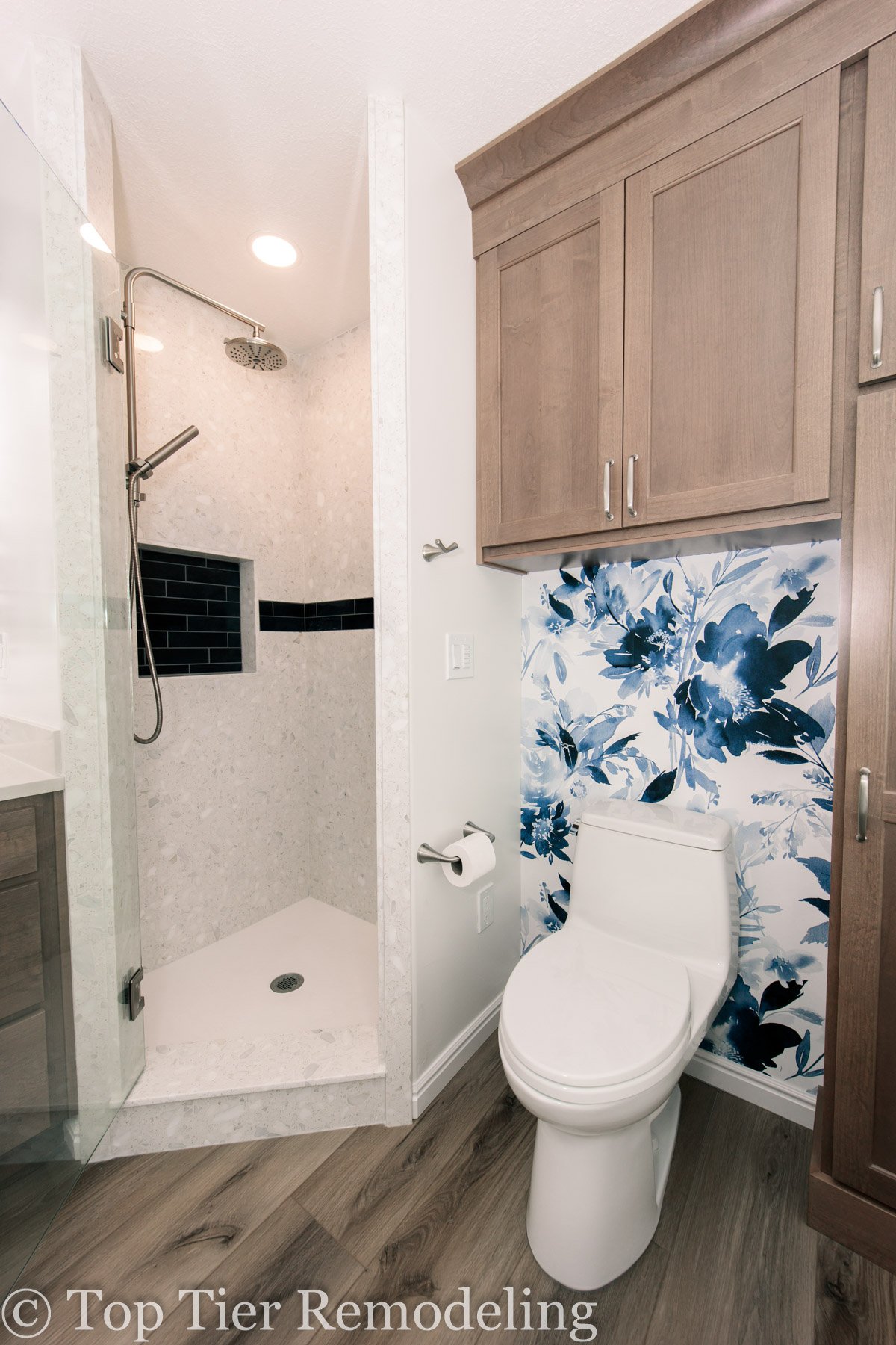Modern bathroom with a walk-in shower featuring a rainfall showerhead, a toilet with a floral blue wallpaper behind it, and wooden cabinets above.