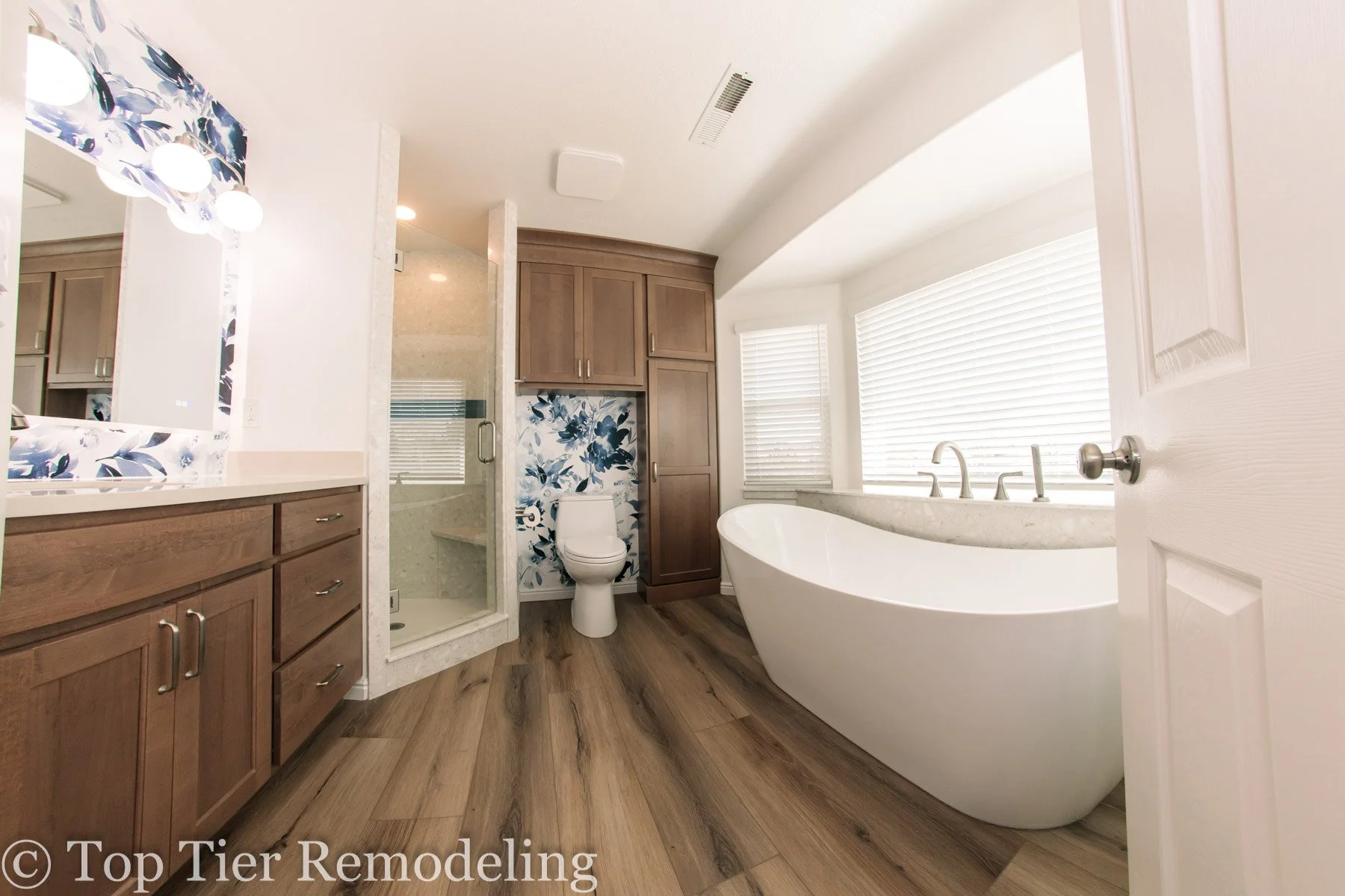 Modern bathroom with free-standing bathtub, large window with blinds, wooden vanity with multiple drawers, framed mirror, ceiling light, shower stall with glass door, white toilet with floral wall behind, wood flooring.