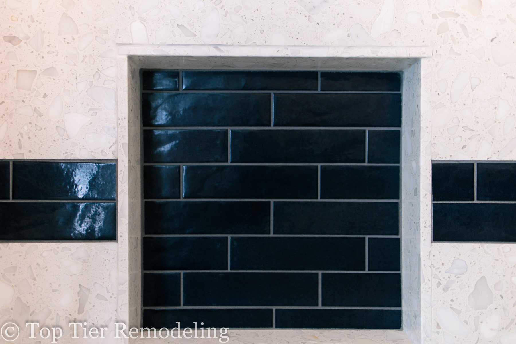 A close-up of a fireplace with a modern design, featuring glossy black brick tiles in a rectangular pattern, surrounded by a white terrazzo wall with small, irregularly shaped pieces.