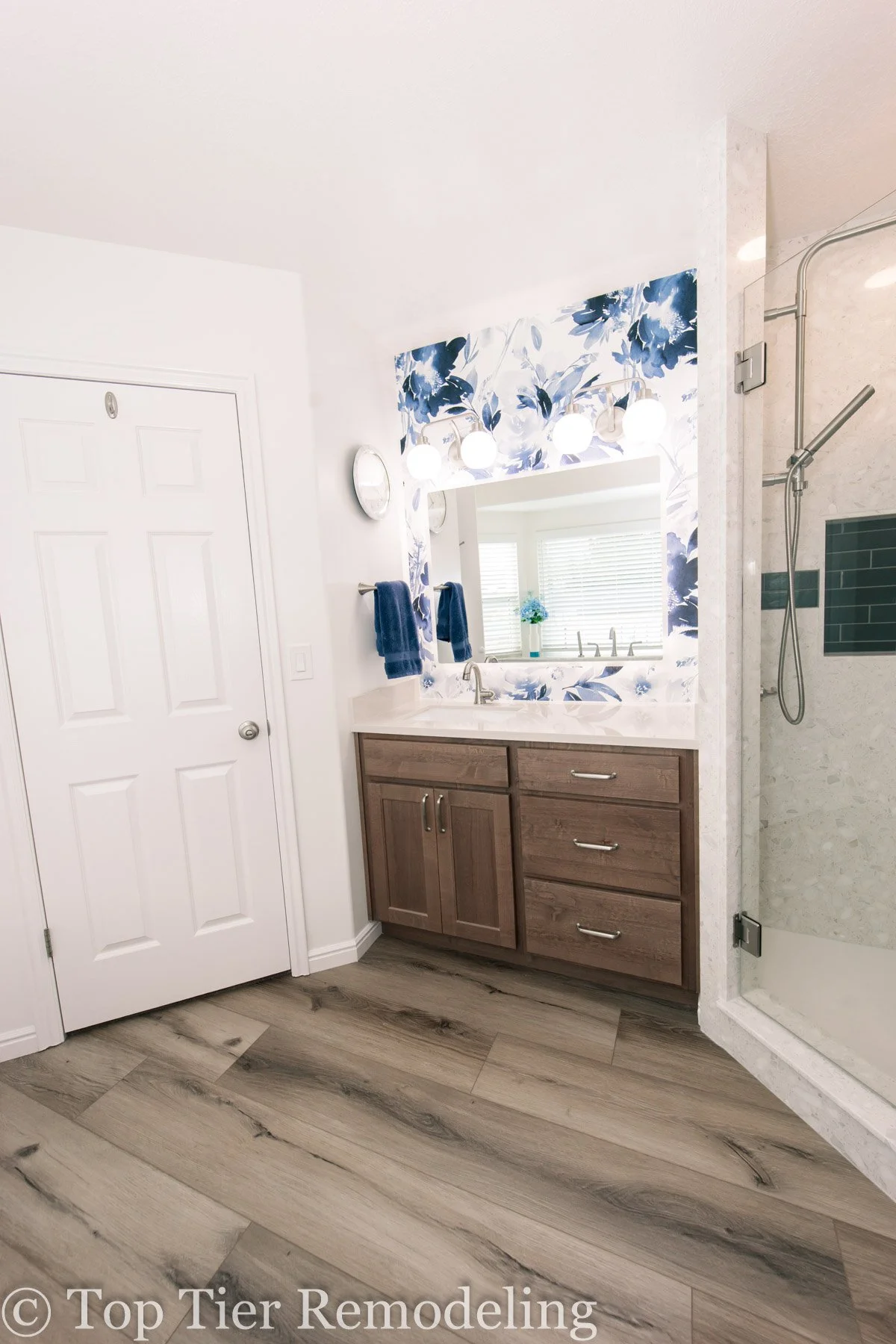 Modern bathroom with wood vanity, large mirror, floral accent wall, white countertop, wall-mounted light fixtures, white door, and walk-in shower with glass door and tile walls.