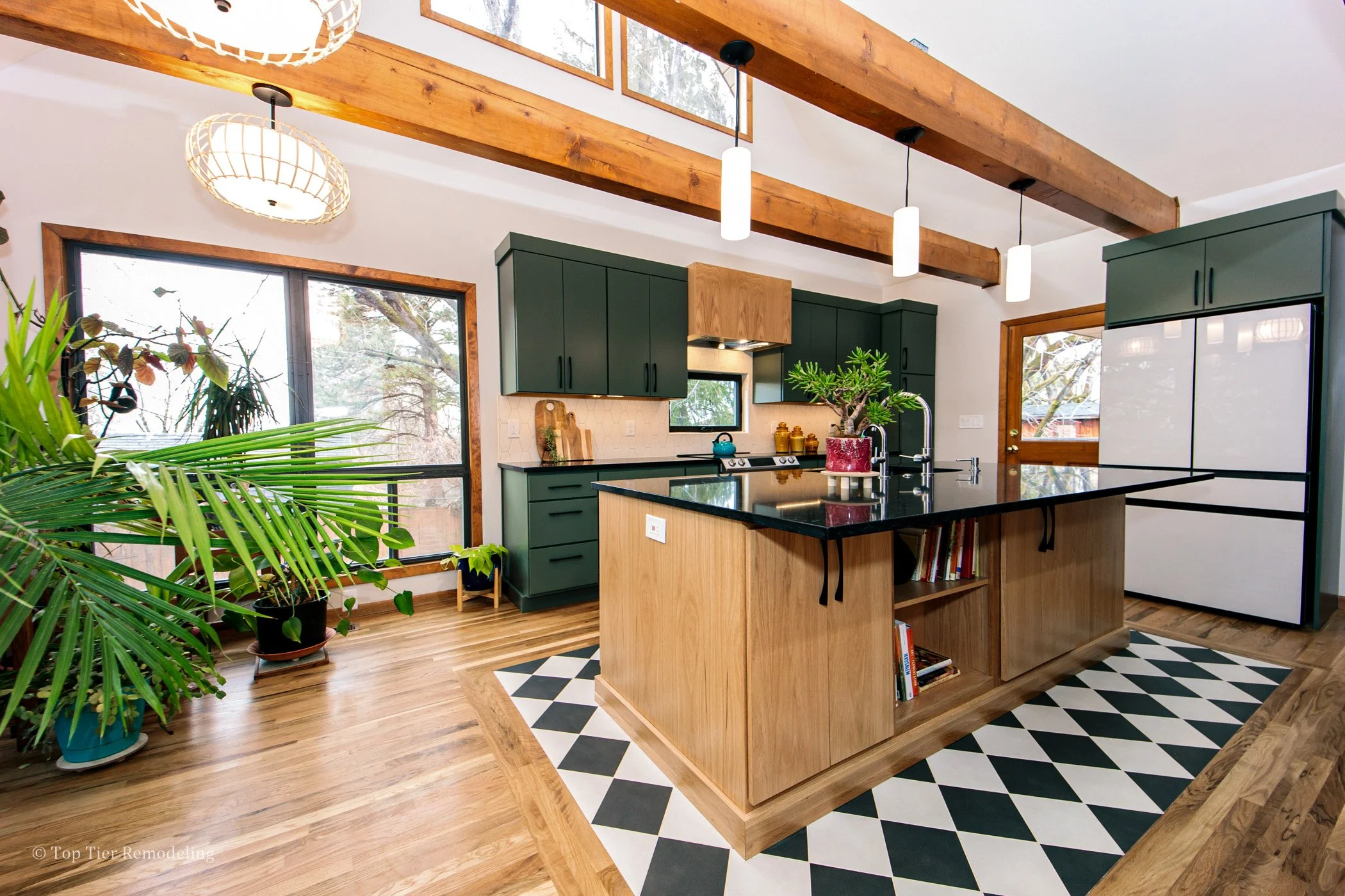 Open kitchen with green cabinets, black countertops, large central island, mixed wood flooring and ceiling beams, and decorative black and white tile pattern near the island. Large windows reveal trees outside, with several potted plants inside.