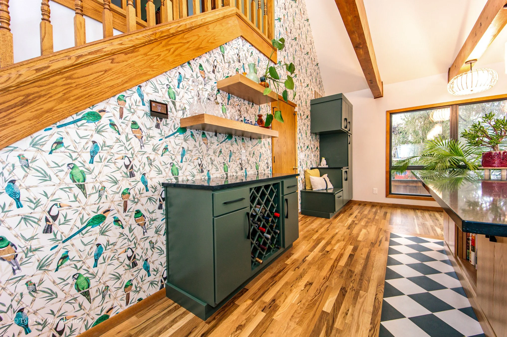 Interior view of a home with a wood staircase, colorful bird-patterned wallpaper, green cabinets, and large windows with plants outside.