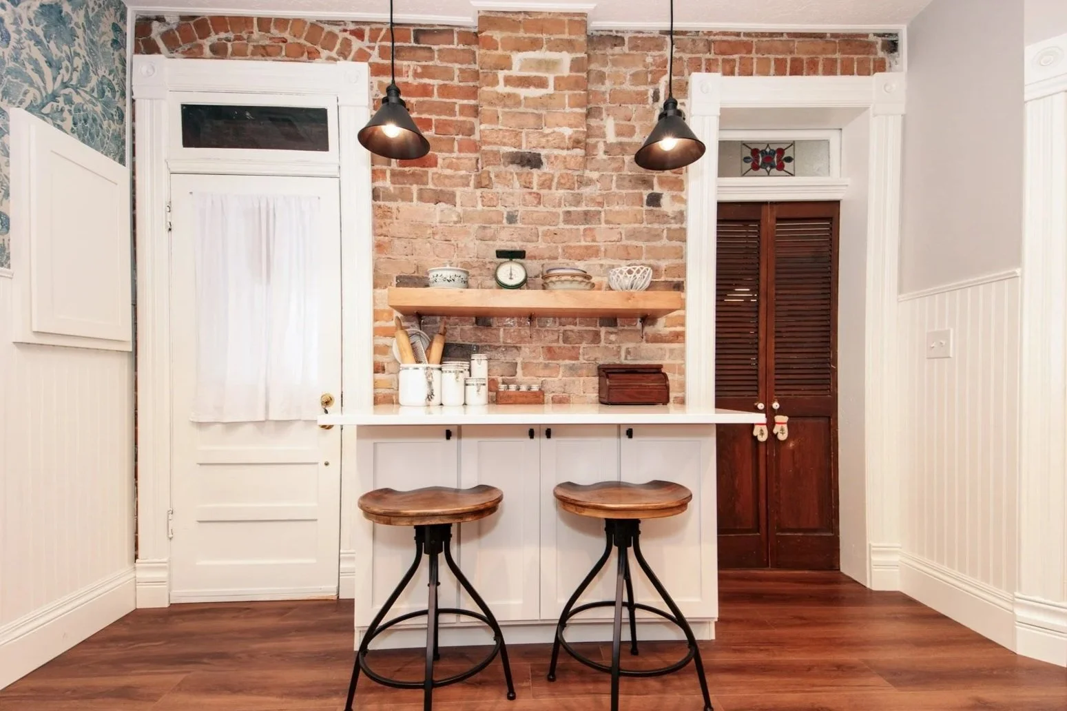 A cozy kitchen corner with a brick accent wall, white cabinets, an open wooden shelf with dishes and a scale, two wood and metal stools, and a brown louvered door.