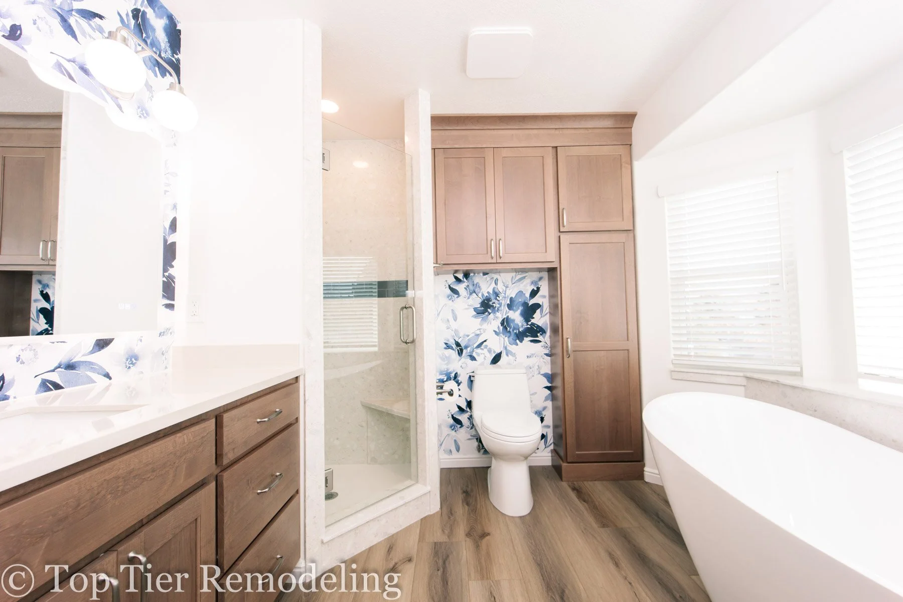 Modern bathroom with wooden cabinets, a white bathtub, a toilet with a blue floral accent wall, a glass shower enclosure, and a window with white blinds.