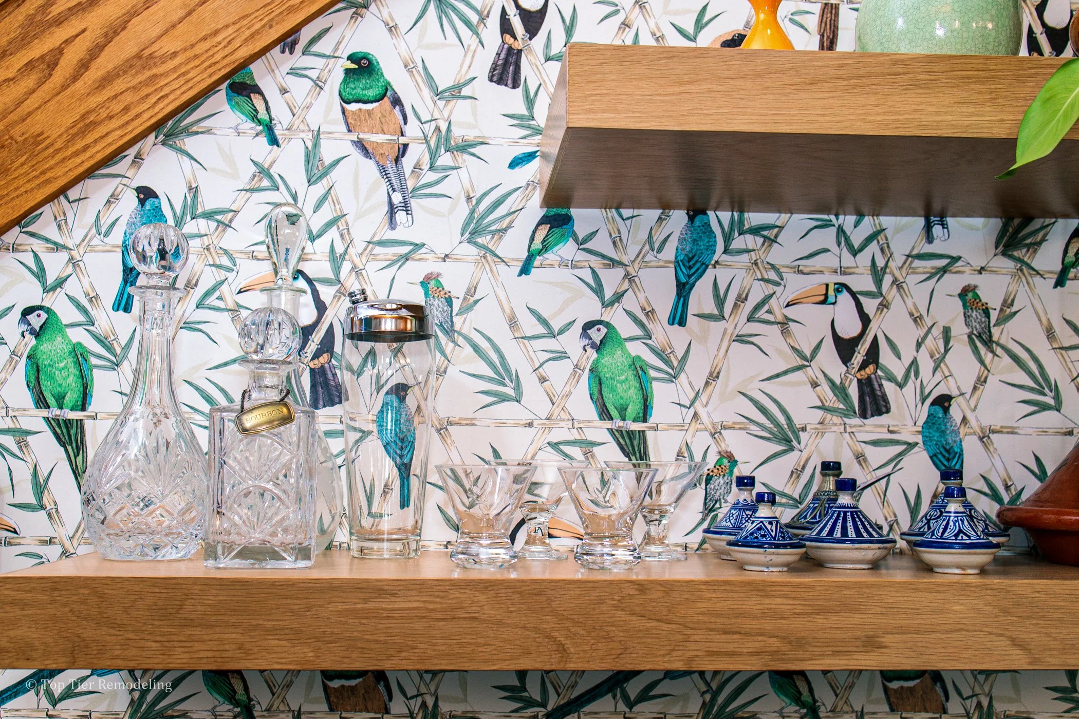 A wooden shelf displaying various clear glass decanters, bowls, and ceramic jars with blue and white designs, set against a tropical bird-patterned wallpaper featuring parrots, toucans, and other exotic birds.