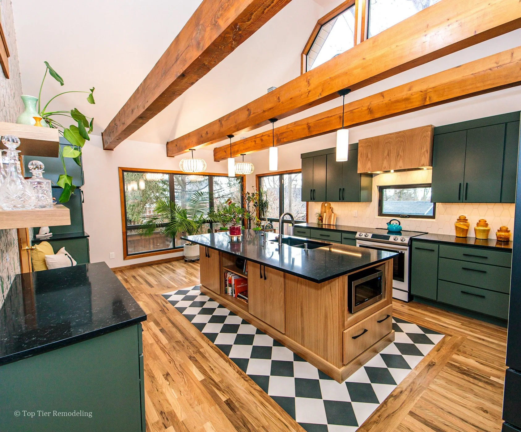 Modern kitchen with wooden beams on vaulted ceiling, black countertops, green cabinets, large windows, and a central kitchen island with open shelving and books. Decor includes potted plants, colorful jars, and a blue teapot. Patterned black and whit