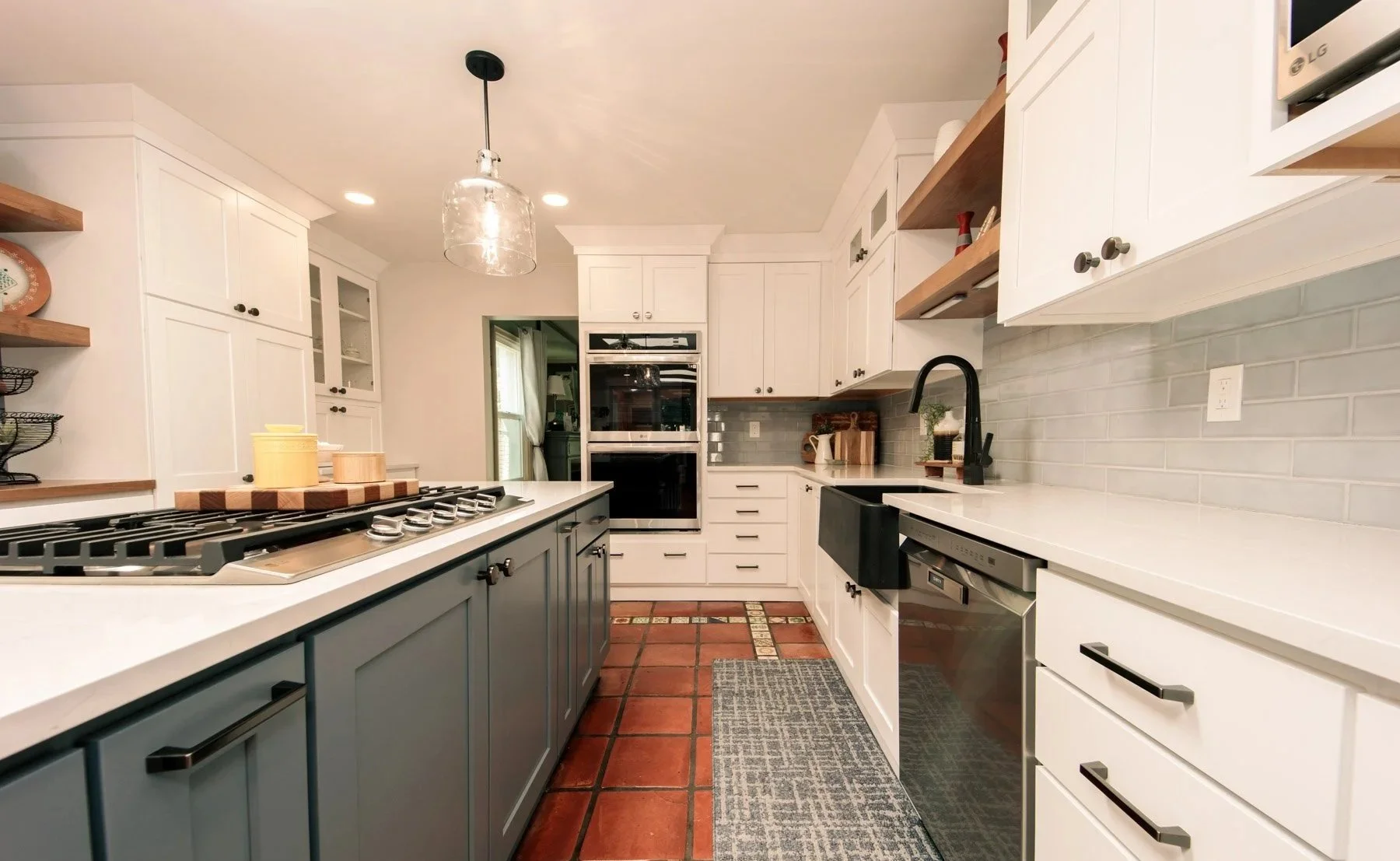 Modern kitchen with white cabinets, black accents, and a gray island. Brick flooring, open shelving, and a glass pendant light. Appliances include a double oven and dishwasher.