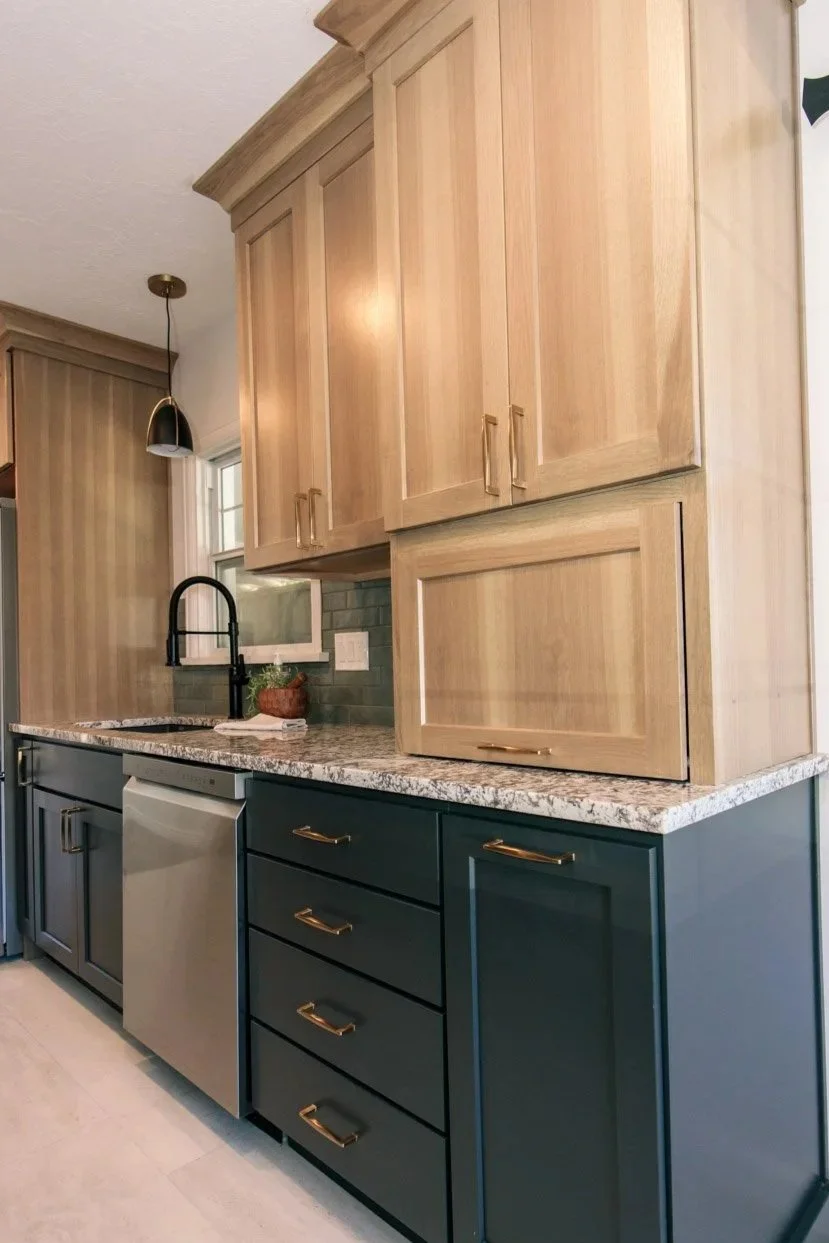 Kitchen with natural wood upper cabinets, dark green lower cabinets with gold handles, granite countertop, black faucet, small window, and a dishwasher.