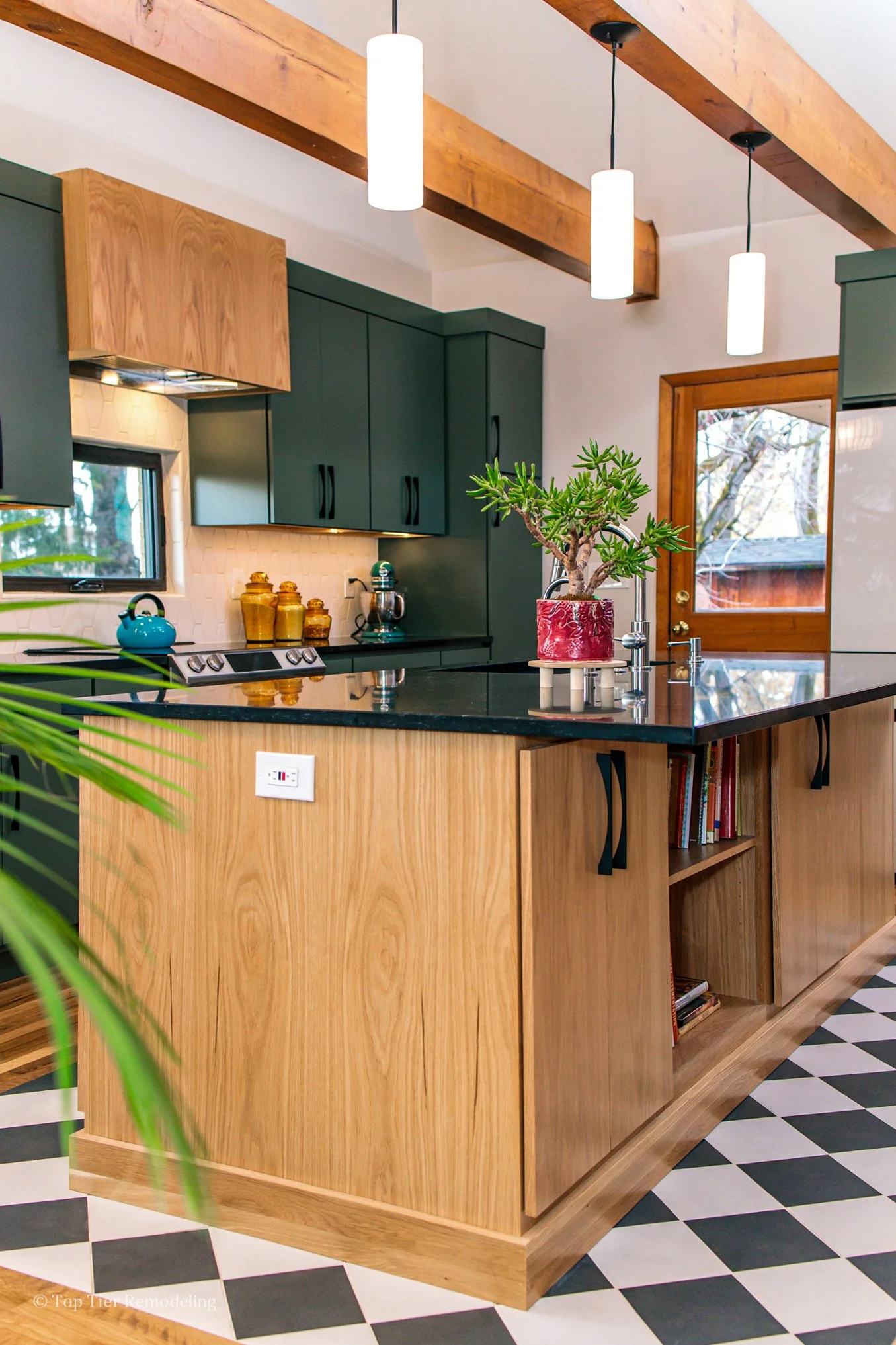 A kitchen with wooden cabinetry painted dark green, a black countertop, and a black and white checkered tile floor. There is a potted succulent plant on the island, a small window, and a door with a window showing outdoor trees. Three pendant lights 