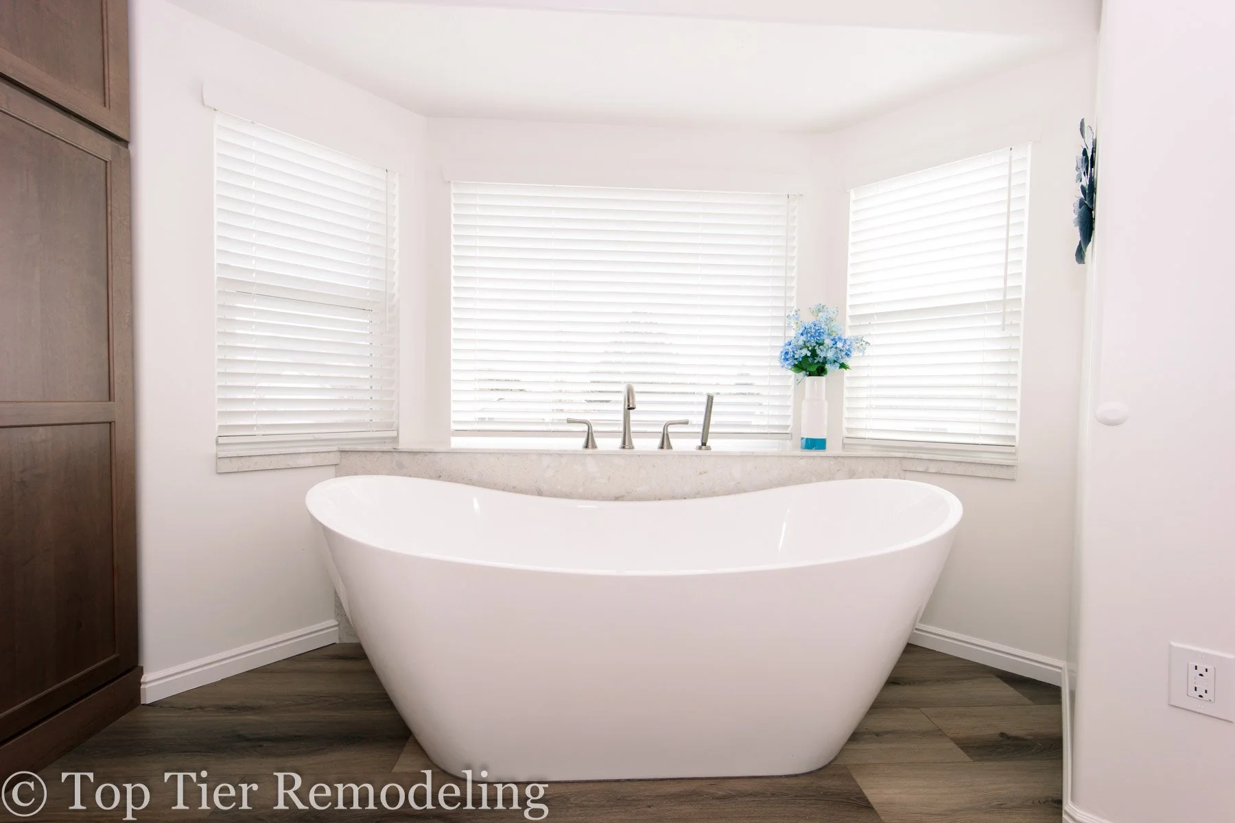 Modern bathroom with a white freestanding bathtub in front of three large windows with white blinds, a flower vase with blue flowers on the ledge, and a wooden cabinet to the left.