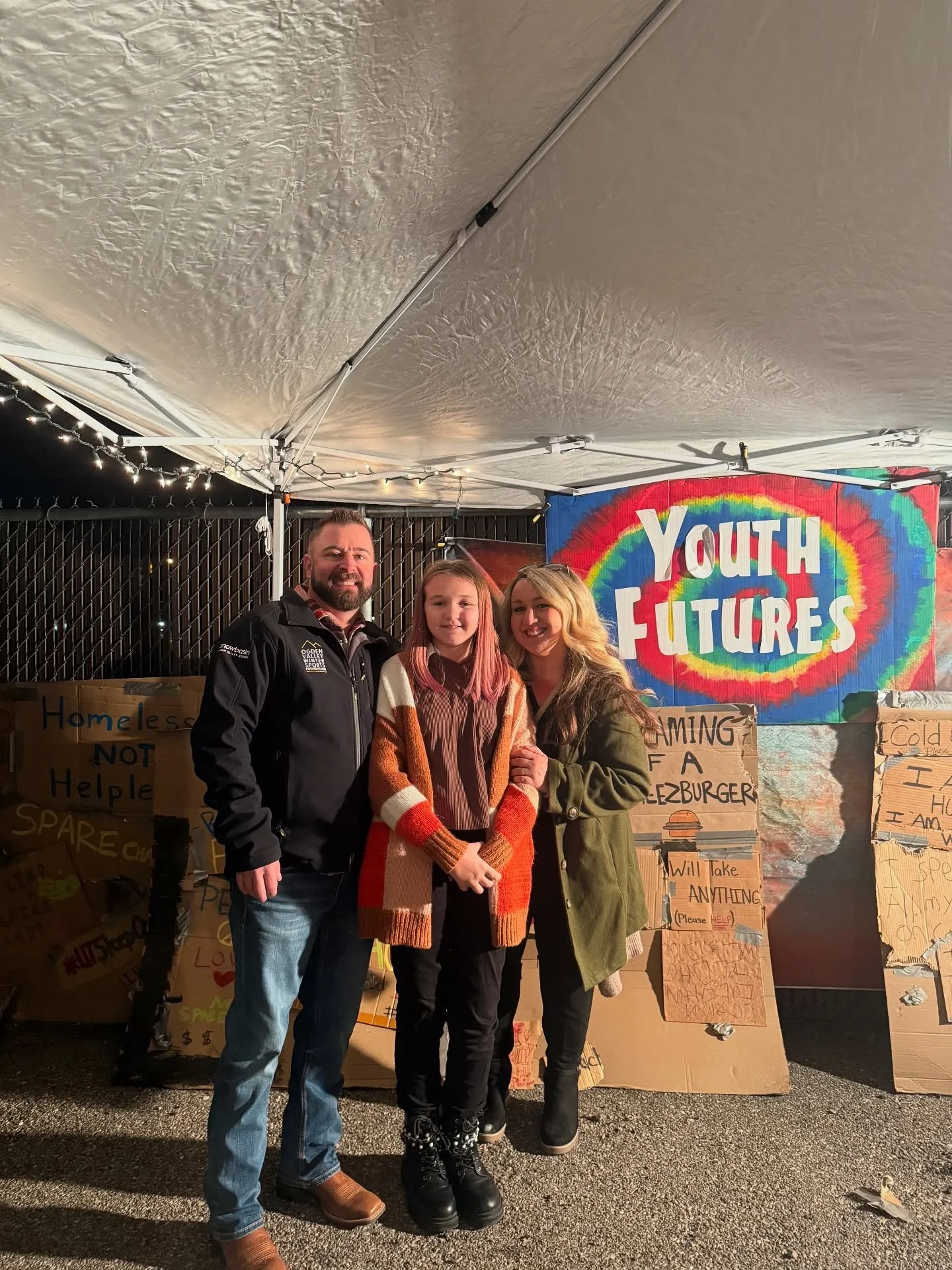 Three people, two women and one girl, standing under a tent at a night event with a 'Youth Futures' rainbow sign, surrounded by cardboard signs with messages.