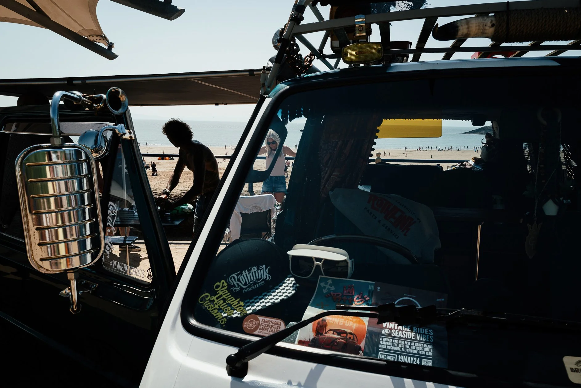 The photo shows the front part of a black and white vehicle, likely a van, with various stickers on the windshield. Behind the vehicle, there is a beach scene with people walking along the shore and the ocean visible in the background. Inside the veh