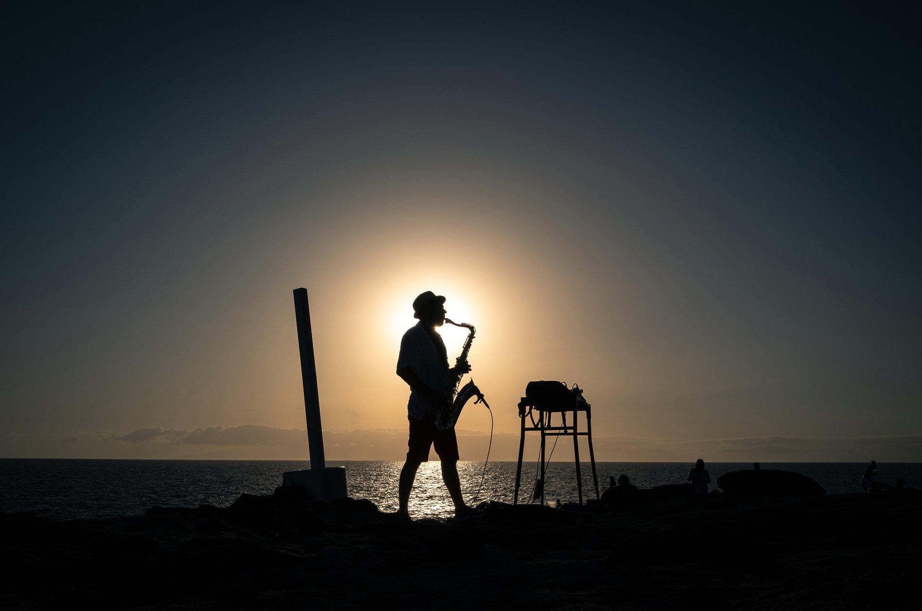 Silhouette of a man with a saxophone playing on the beach at sunset, near musical equipment, with the ocean and setting sun in the background.
