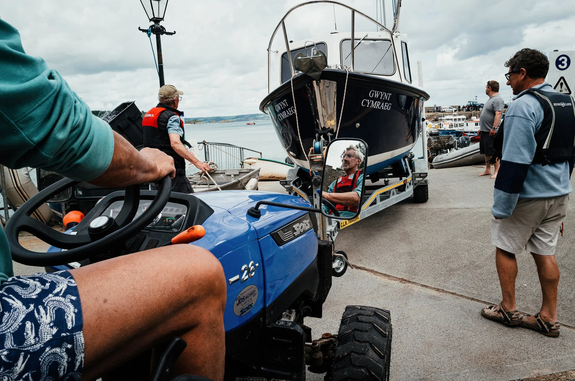 A boat is being towed on a trailer by a blue golf cart at a marina. Several people are present, some wearing life jackets, with water and other boats visible in the background on a cloudy day.