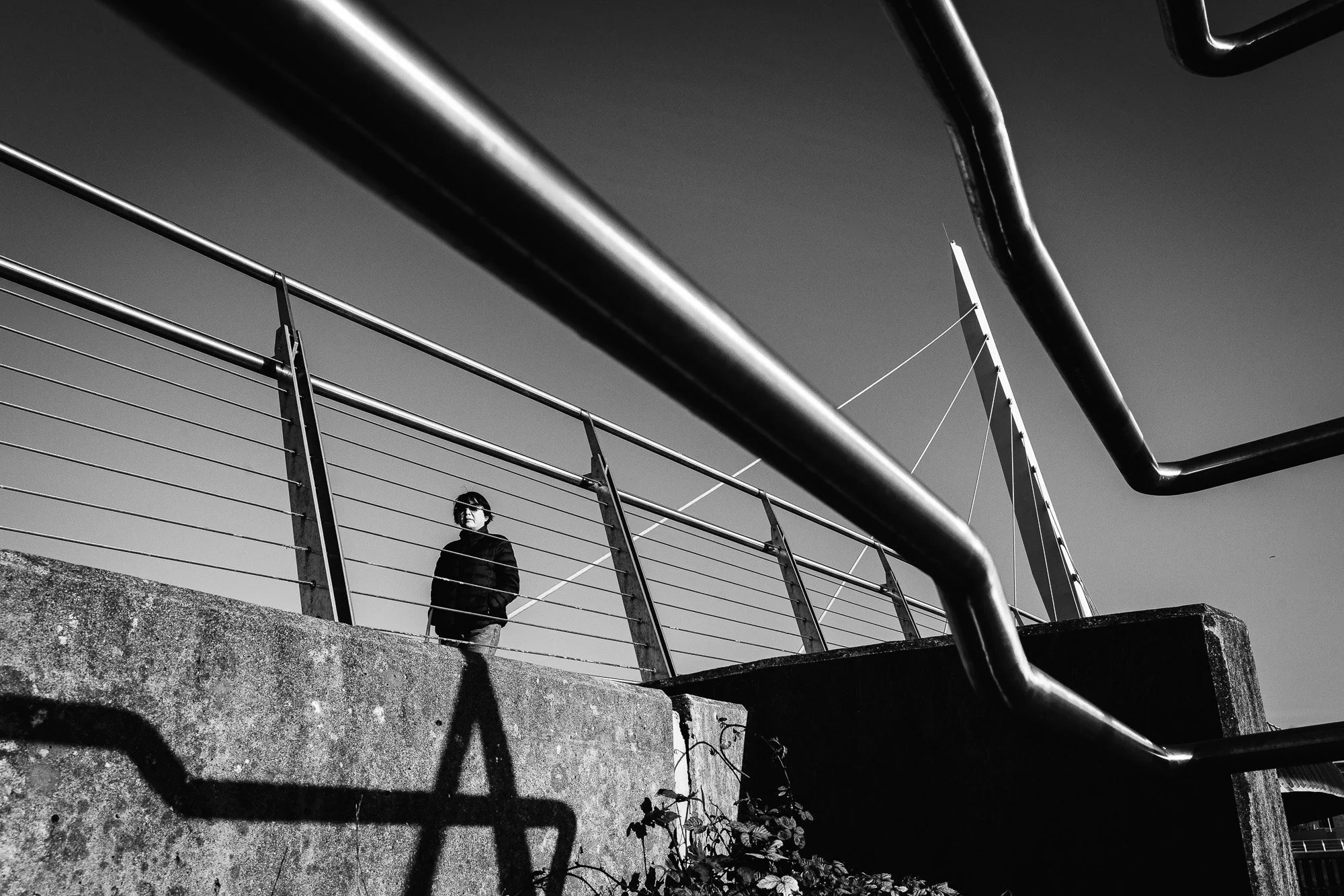 A person standing on concrete steps beneath a metal railing, with large metal pipes crossing above and a pole with cables in the background. The image is in black and white, with shadows cast on the steps.