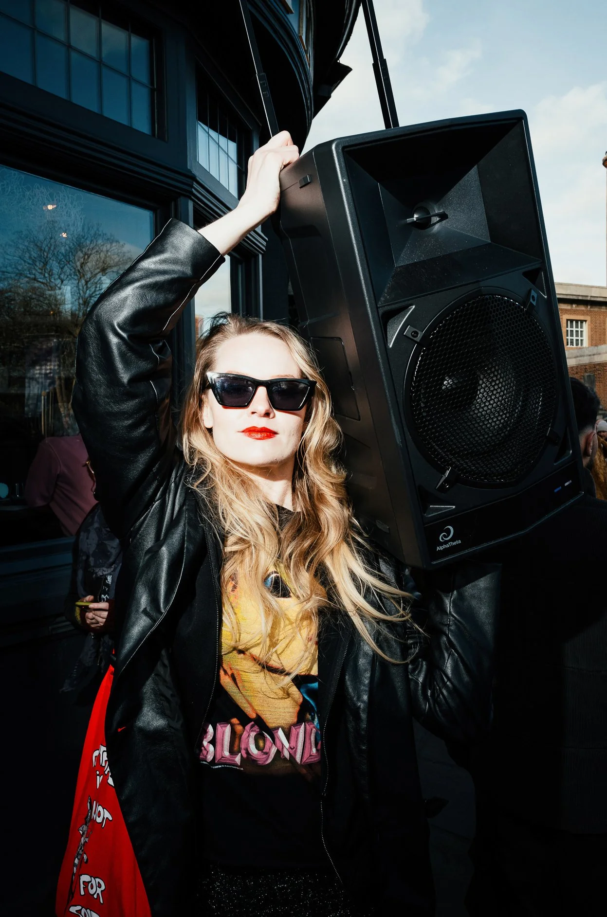 A woman with blonde hair, wearing sunglasses, a black leather jacket, and bright red lipstick, holding a large speaker on her shoulder outdoors during daytime.