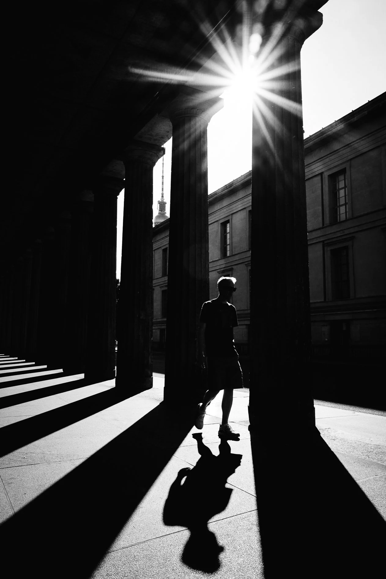 A black and white photo of a person walking under classical columns, with sunlight shining through and casting shadows on the ground.