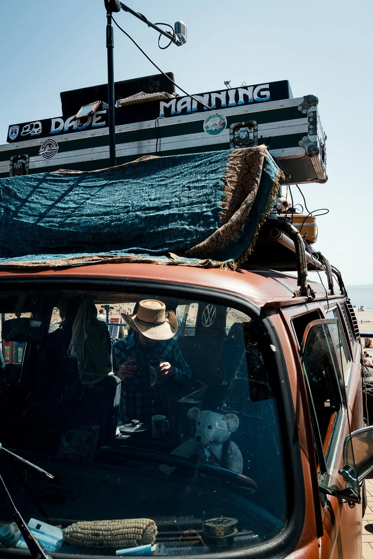 A vintage orange Volkswagen van parked at the beach with a rack on top holding various items including a banner that reads 'Dude Maning' and a small sticker with a mountain scene. The van has a reflection in the windshield showing a person wearing a 