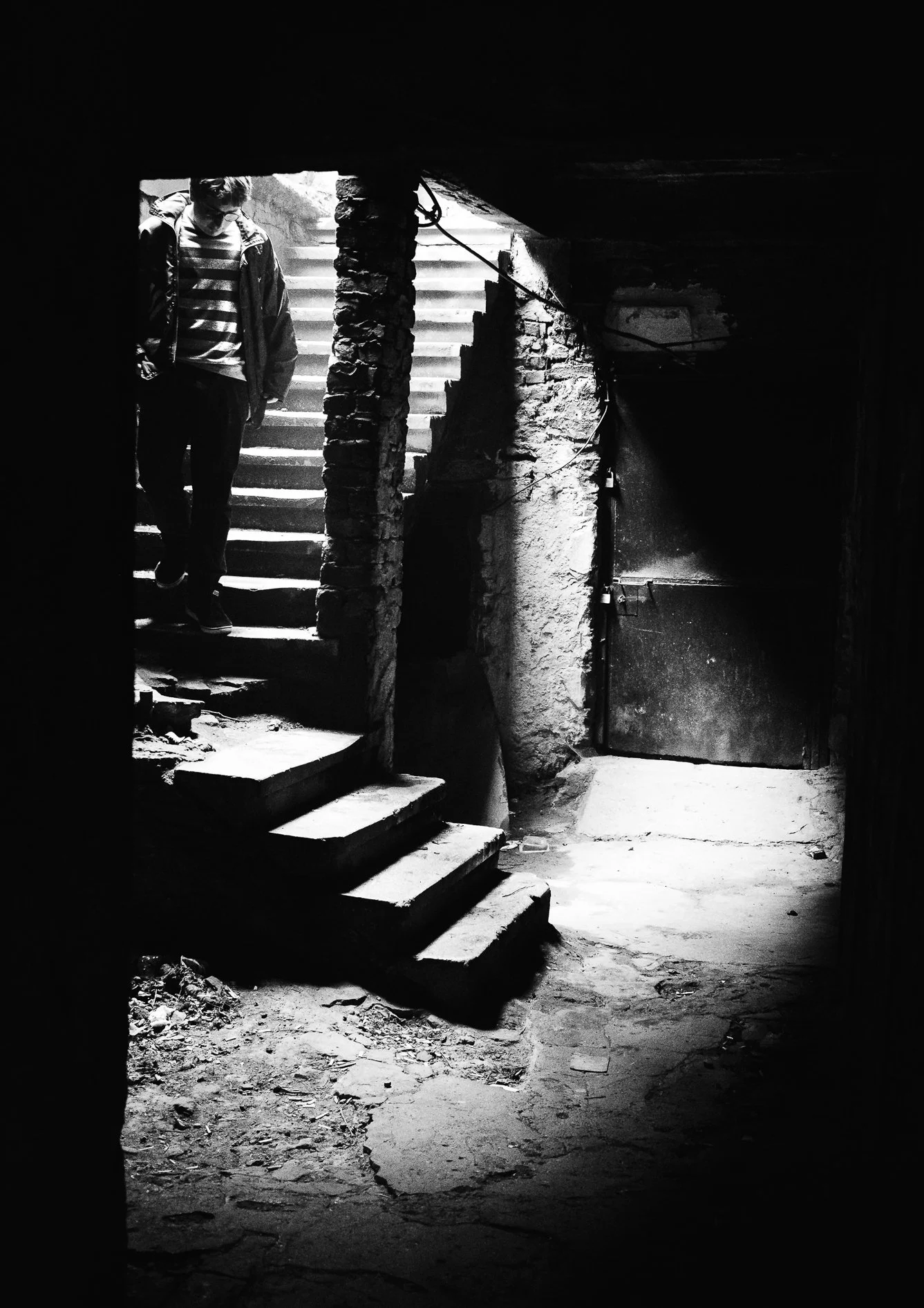 A black and white photo of a person descending worn stone stairs in a dark, possibly basement or cellar, area with rough textured brick and concrete walls.