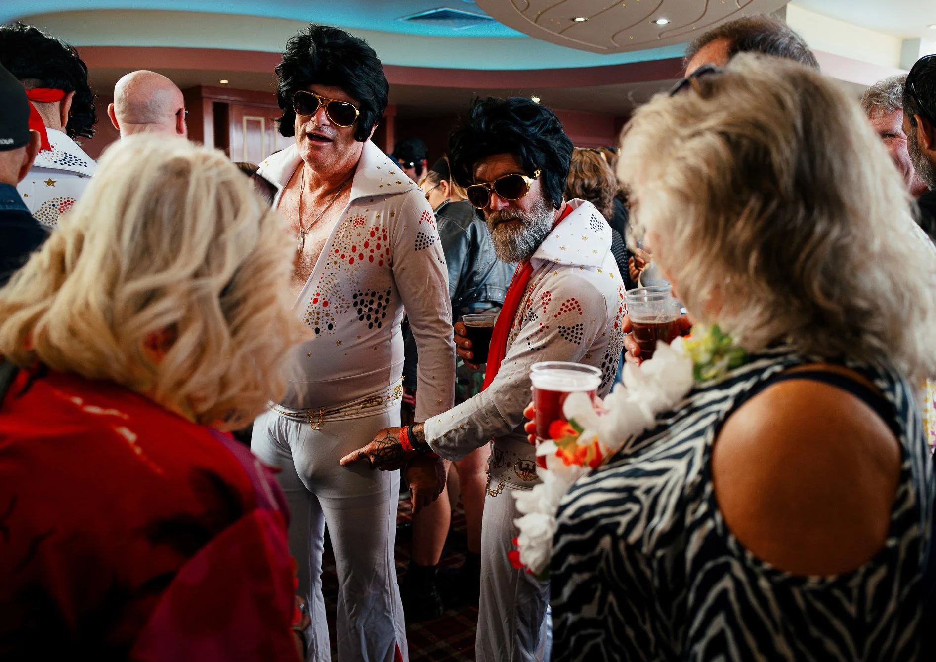 People at a party dressed in 1970s style outfits, with some wearing sunglasses, holding drinks, and engaging in conversation.