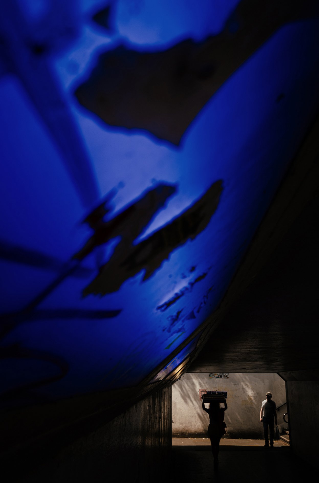 People walking under a bridge with a blue-lit sign overhead, one person carrying a box on their head, another walking alongside.