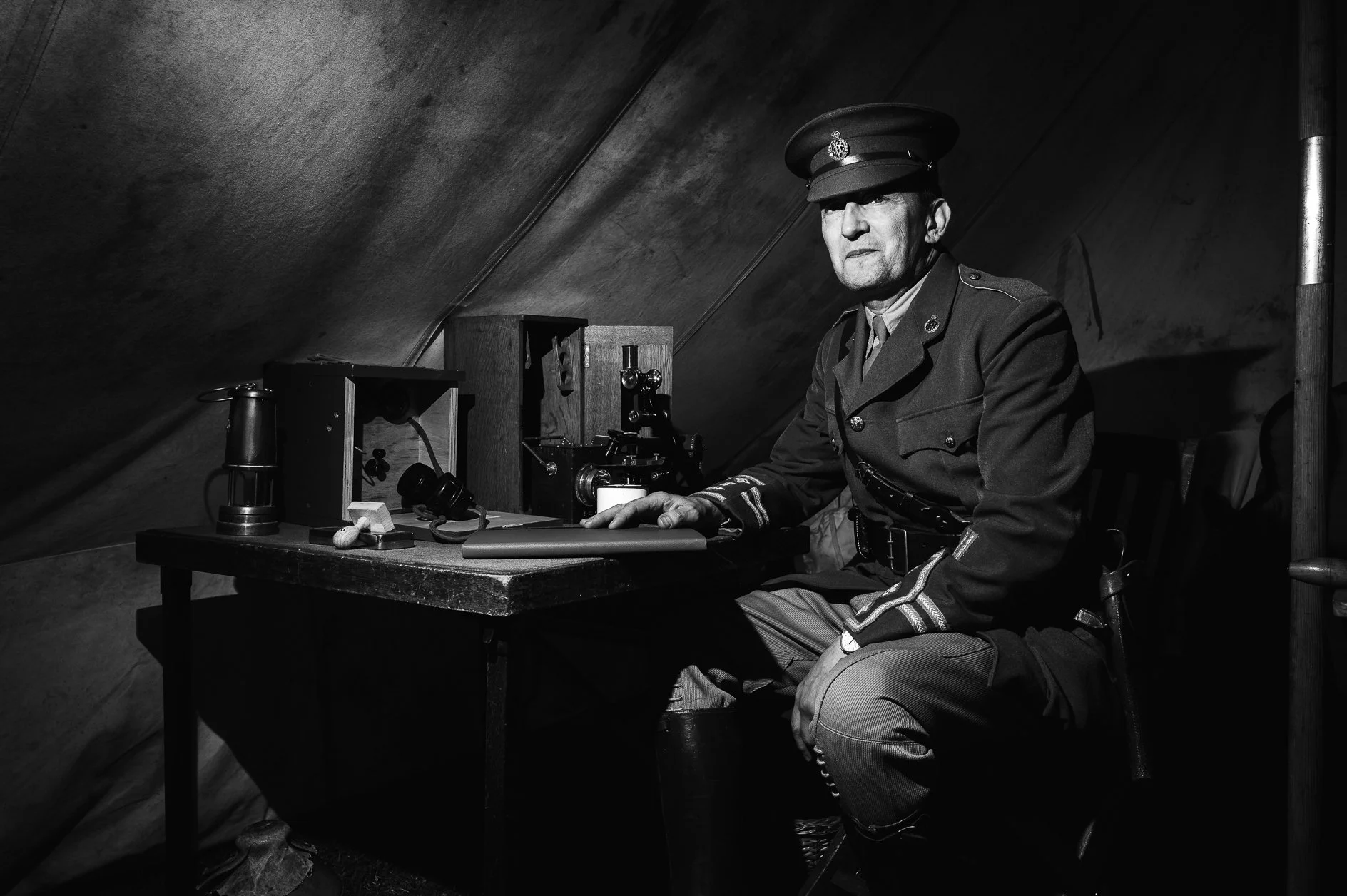 Black and white photo of a man in a military uniform sitting at a desk inside a tent. He is wearing a military cap and looking directly at the camera. The desk has various objects, including a telephone, a small box, and a mug. The background shows a