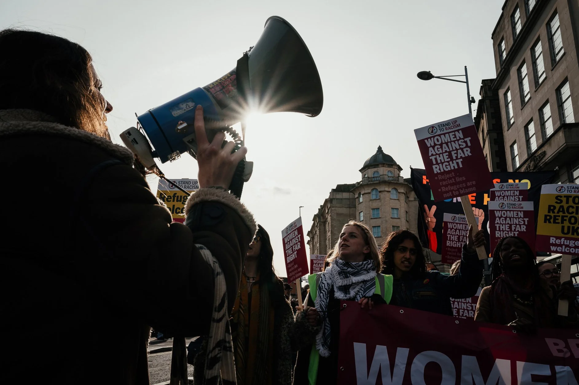 Protesters holding signs that say "Women Against the Far Right" and "Stop Racism" during a rally on a city street, with one woman speaking into a megaphone, and tall buildings and a streetlamp in the background.