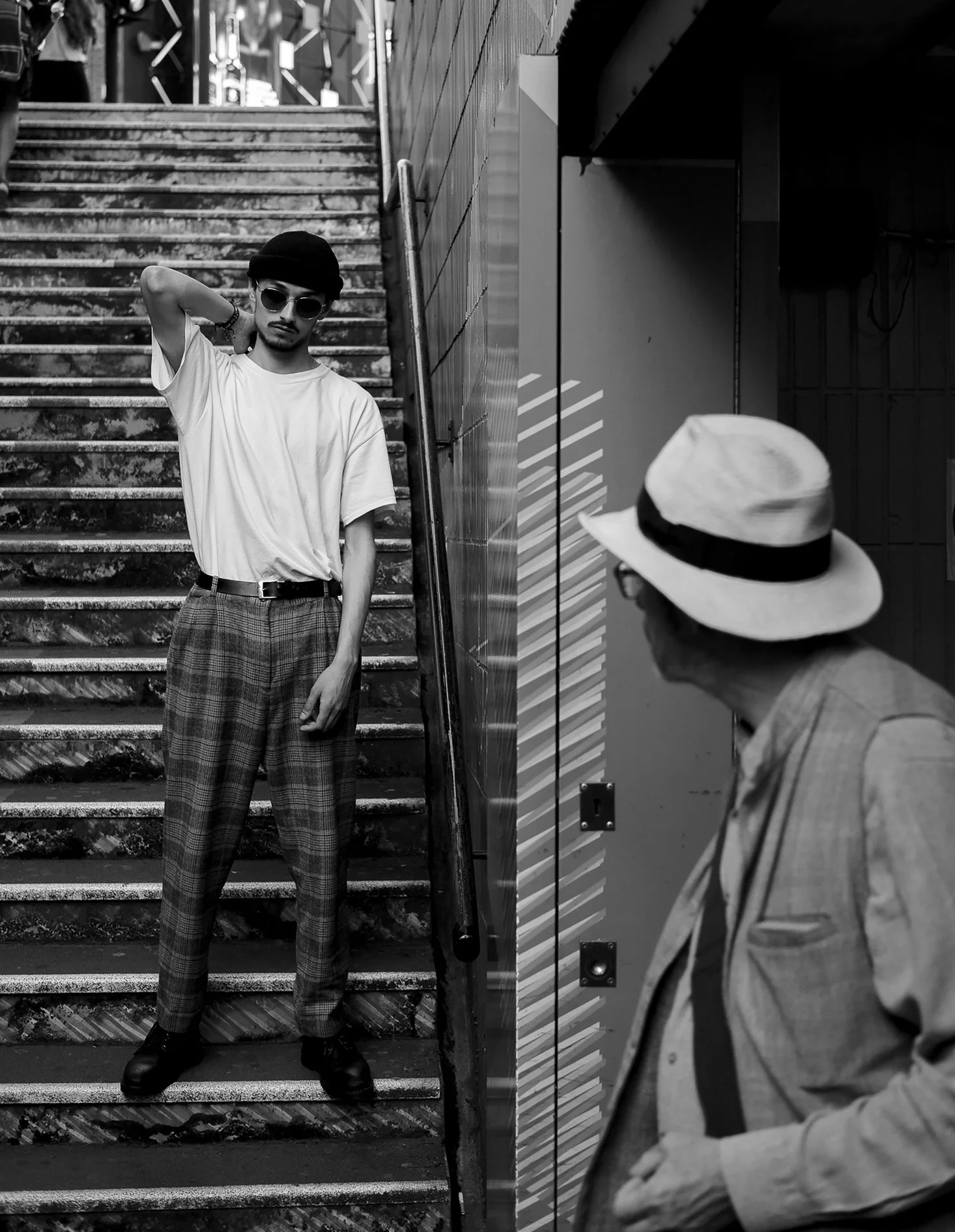 A black and white photo of a young man standing on the stairs with his hand behind his head, wearing sunglasses, a loose white t-shirt, plaid trousers, and a cap, with another person in the foreground wearing a hat.