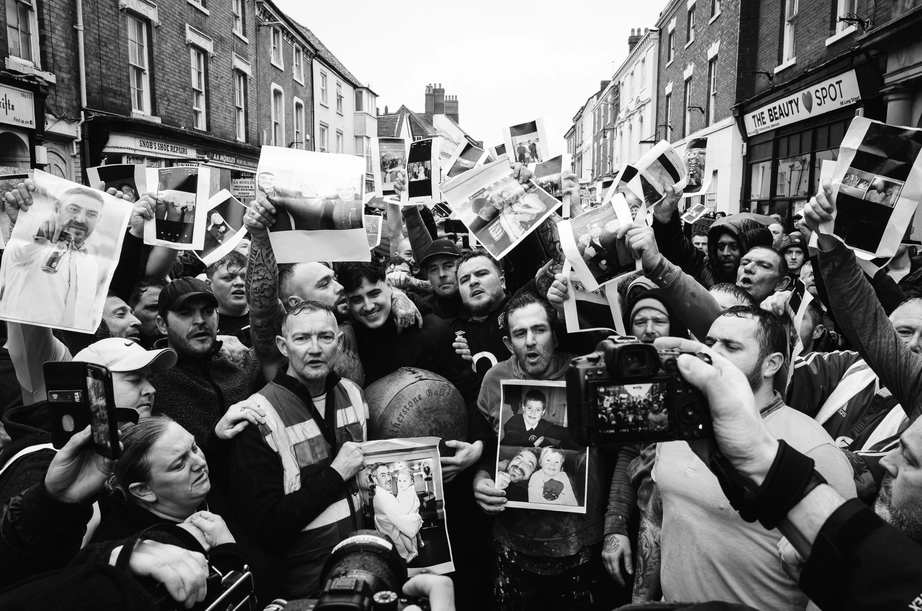 A black-and-white photo of a large crowd of people gathered on a city street, holding photos and signs, some people wearing jackets. The street is lined with buildings with storefronts, including a shoe repair shop and a beauty spot. The crowd appear