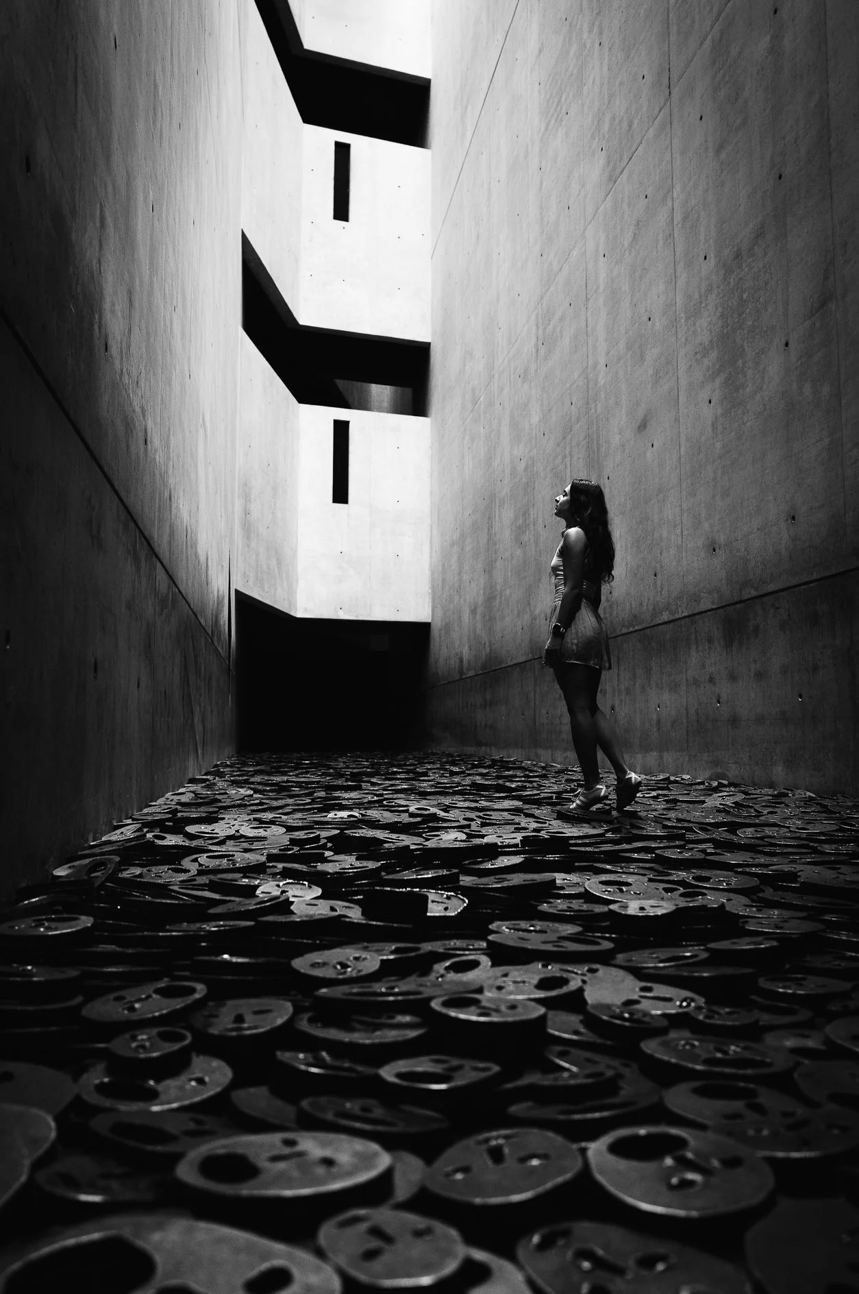 A woman with long hair and a sleeveless dress walking in an indoor space with high concrete walls, patterned floor, and light coming from above, in black and white.