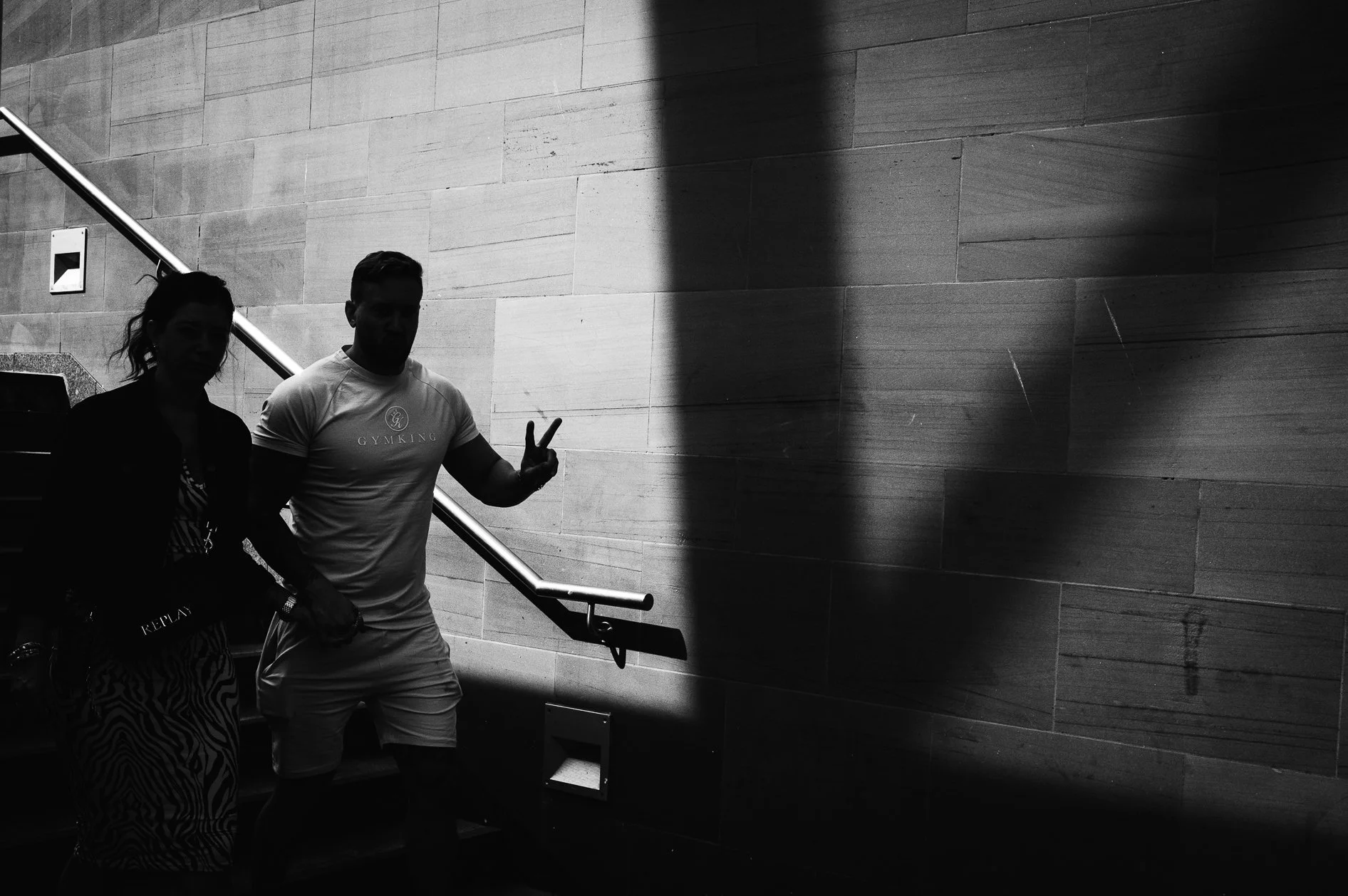 Two people standing on stairs against a brick wall, one making a peace sign, both silhouetted due to lighting.